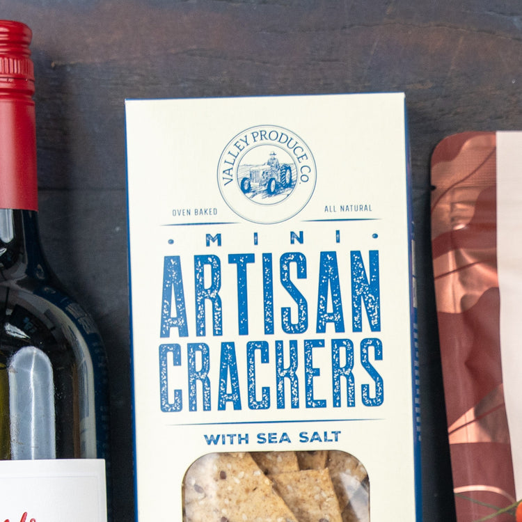 A close-up of The Hamper Boutique Co Savoury Selection Gift Hamper beside a bottle of red wine and part of another package on a dark surface.