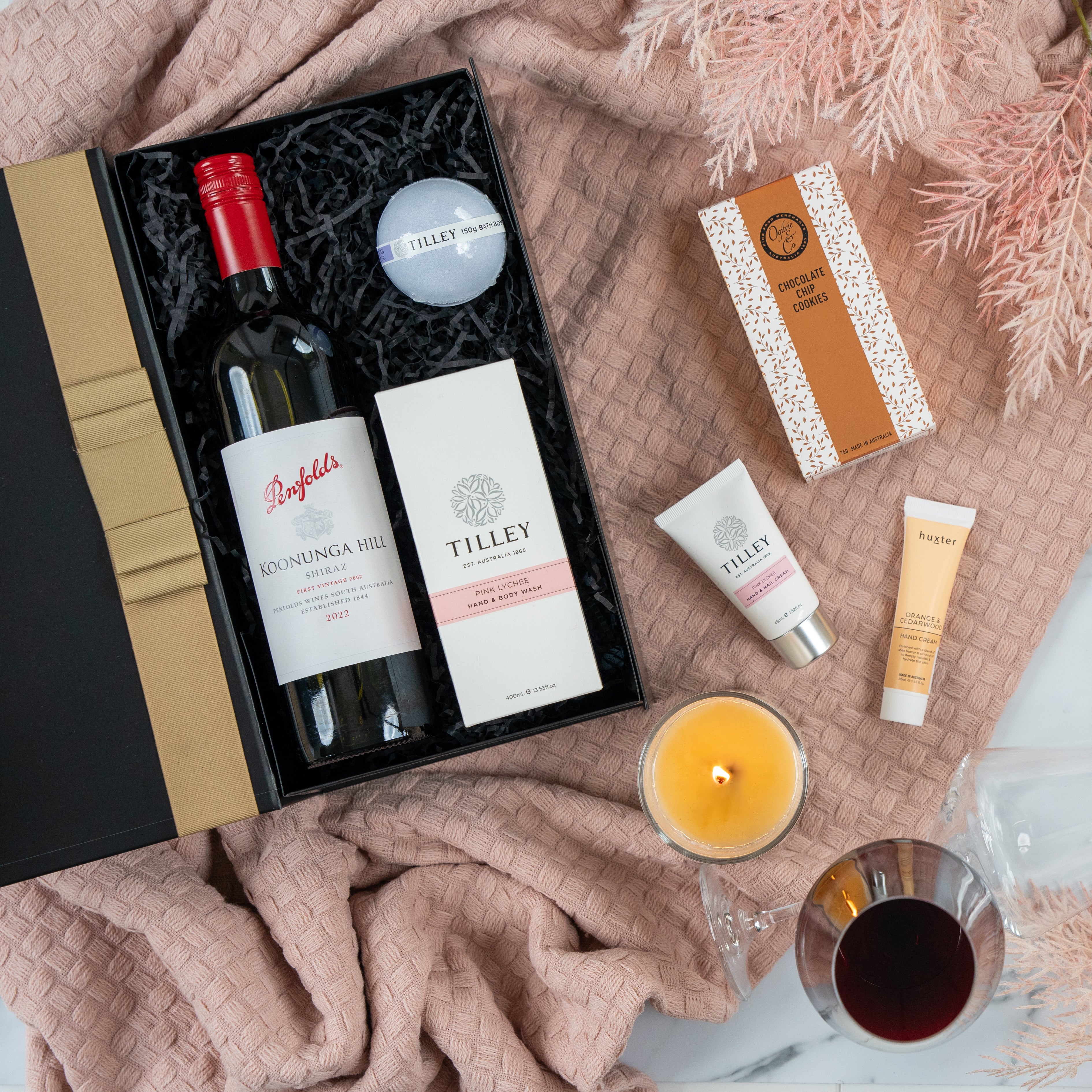 The Hamper Boutique Co’s Pamper and Indulge Gift Hamper includes Penfolds wine, Tilley soap and hand cream, chocolate, a candle, and skincare items—perfect for relaxing with red wine on a pink textured blanket.