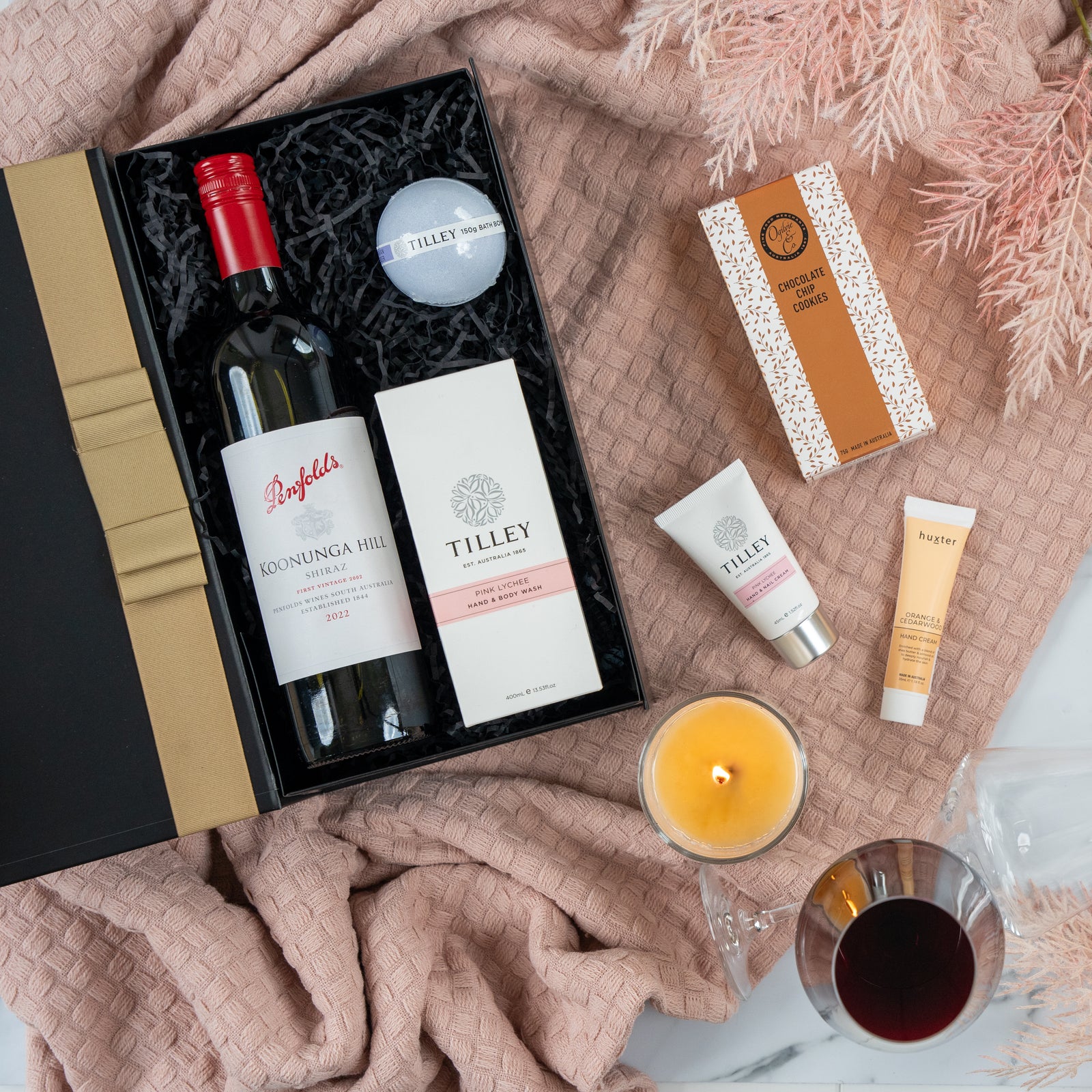 The Hamper Boutique Co’s Pamper and Indulge Gift Hamper includes Penfolds wine, Tilley soap and hand cream, chocolate, a candle, and skincare items—perfect for relaxing with red wine on a pink textured blanket.