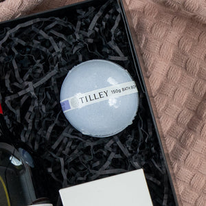 The Hamper Boutique Co Pamper and Indulge Gift Hamper features a Tilley 150g bath bomb on black crinkled paper in a gift box, beside a bottle, white box, and a textured pink towel.