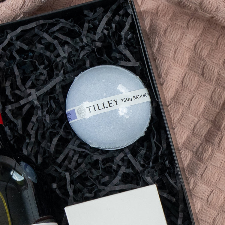 The Hamper Boutique Co Pamper and Indulge Gift Hamper features a Tilley 150g bath bomb on black crinkled paper in a gift box, beside a bottle, white box, and a textured pink towel.