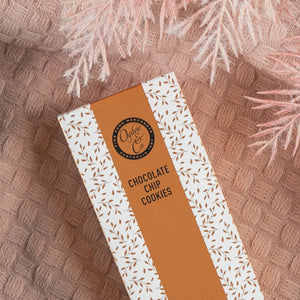 A rectangular box of The Hamper Boutique Co Pamper and Indulge Gift Hamper with a white and brown leaf pattern, displayed on a textured pink surface with soft pink foliage above.