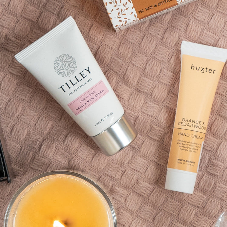 Flat lay of The Hamper Boutique Co Pamper and Indulge Gift Hamper, featuring a cream knit surface, lit candle, Tilley Pink Lychee and Huxter Orange & Cedarwood hand creams, plus a glimpse of a patterned box.