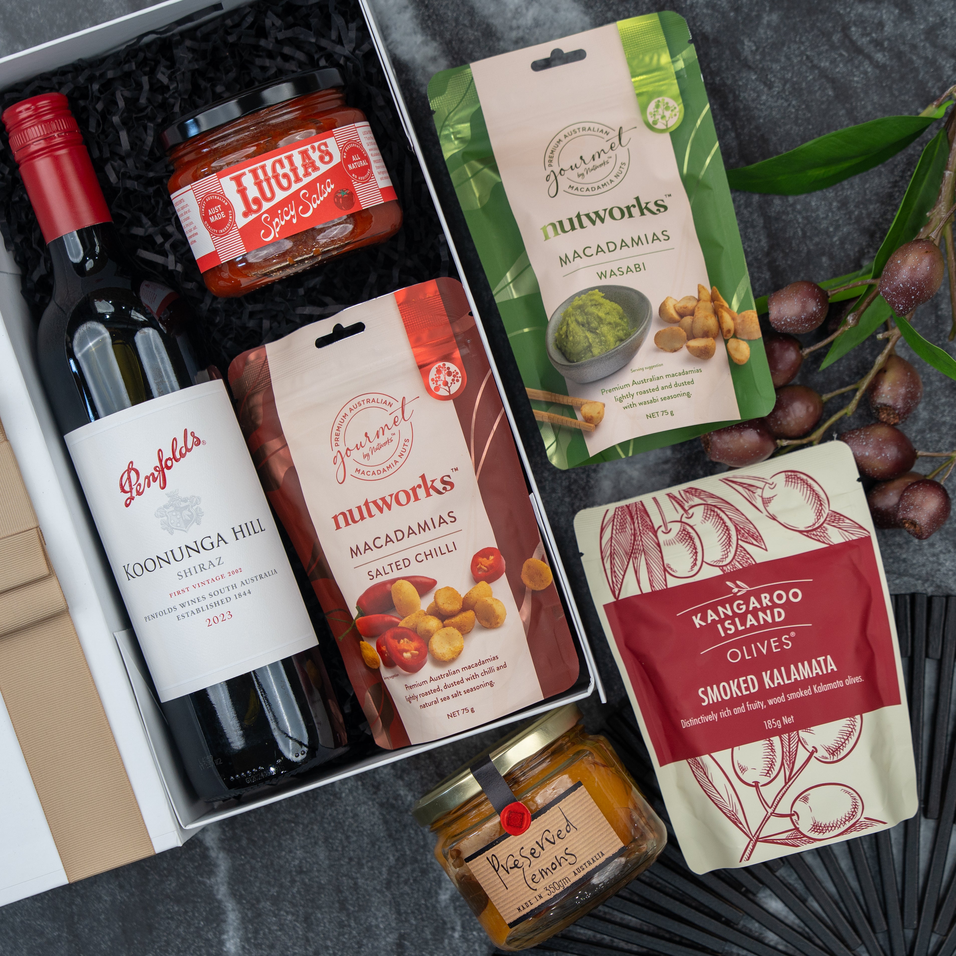 The Hamper Boutique Co’s Flavour Fiesta Gift Hamper features a bottle of Penfolds red wine, Lucia’s spicy salsa, wasabi and salted chili Nutworks macadamias, olives, preserved lemons, and grapes – all arranged on a dark background.