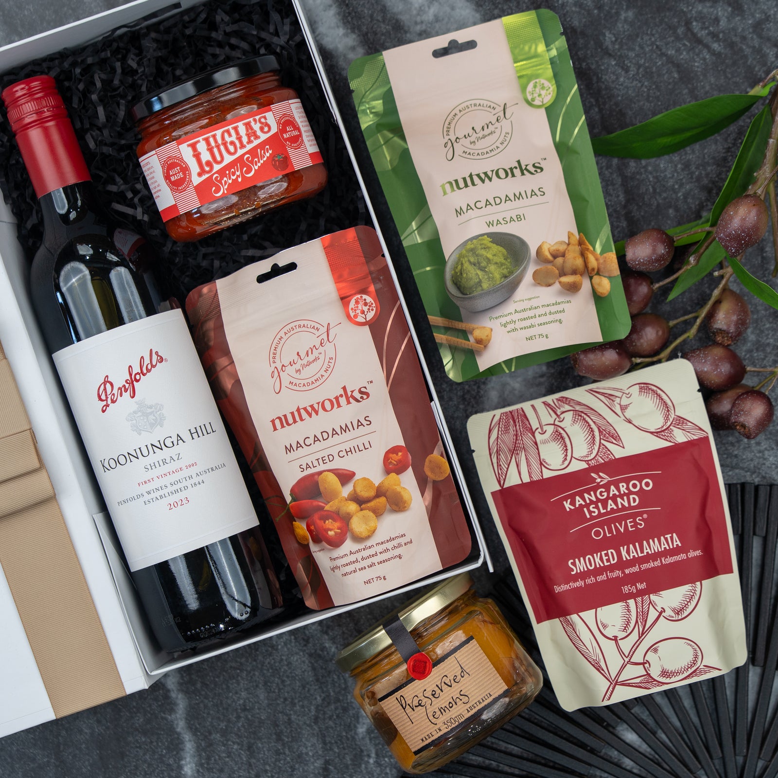 The Hamper Boutique Co’s Flavour Fiesta Gift Hamper features a bottle of Penfolds red wine, Lucia’s spicy salsa, wasabi and salted chili Nutworks macadamias, olives, preserved lemons, and grapes – all arranged on a dark background.