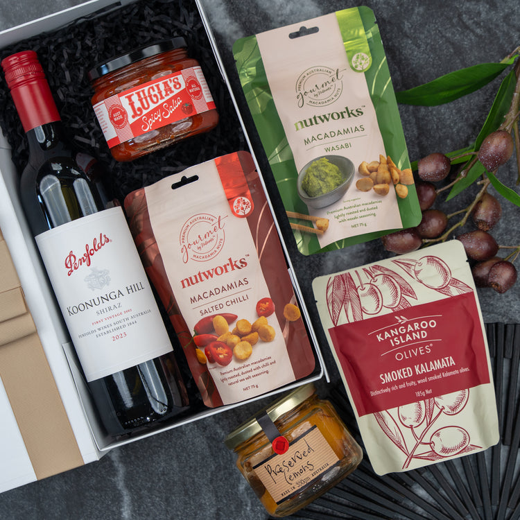 The Hamper Boutique Co’s Flavour Fiesta Gift Hamper features a bottle of Penfolds red wine, Lucia’s spicy salsa, wasabi and salted chili Nutworks macadamias, olives, preserved lemons, and grapes – all arranged on a dark background.
