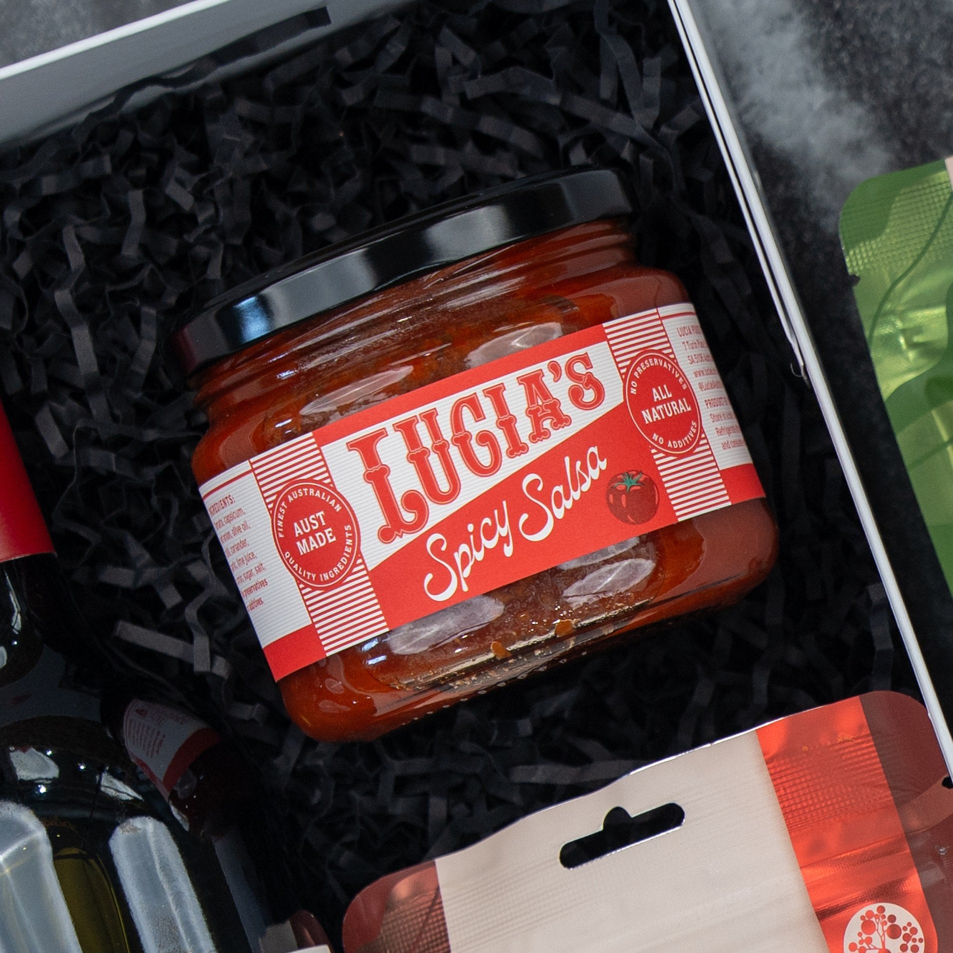 A jar of Lucia’s Spicy Salsa rests on black crinkle paper in The Hamper Boutique Co’s Flavour Fiesta Gift Hamper, surrounded by assorted packaged treats.