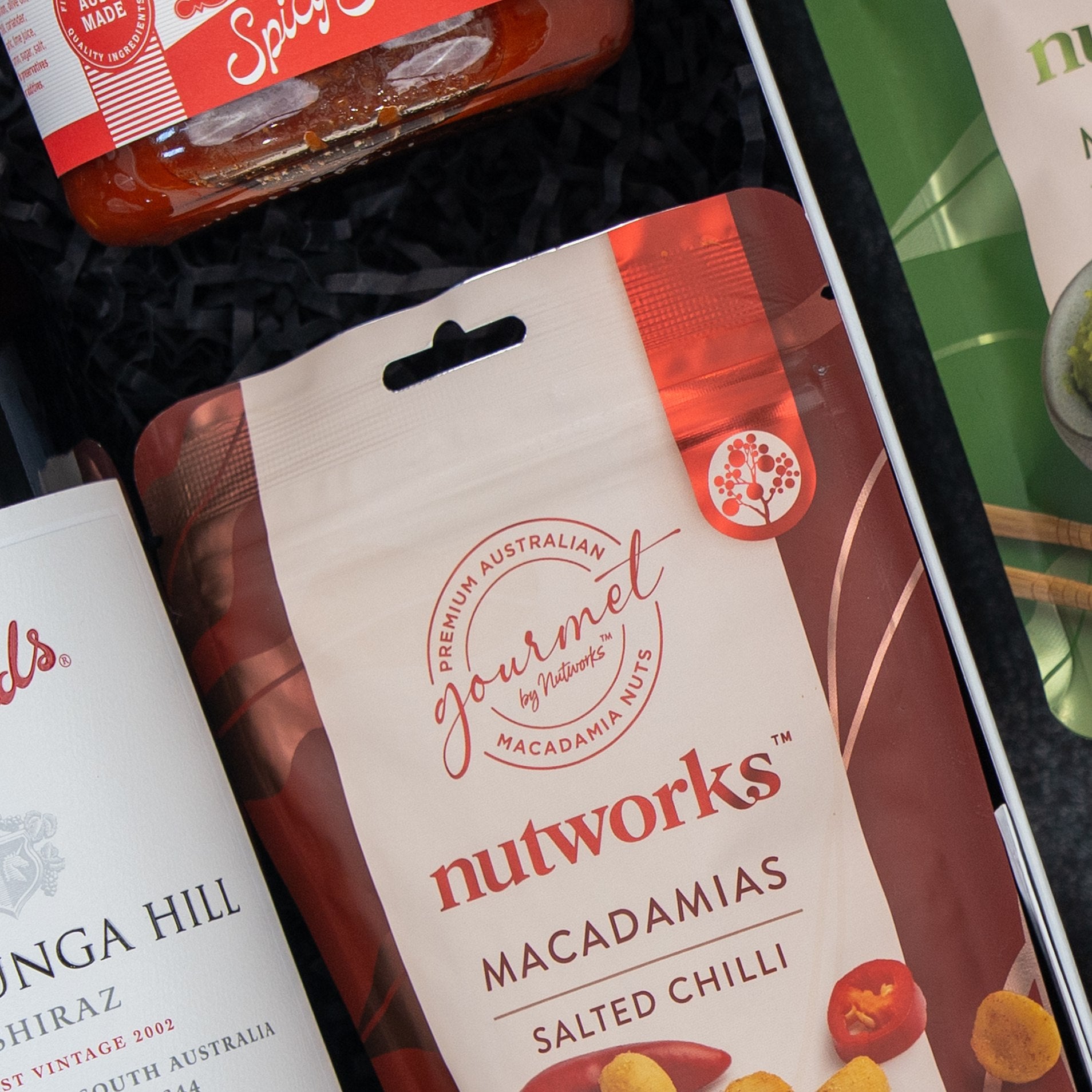 The Hamper Boutique Co Flavour Fiesta Gift Hamper includes spicy tomato chutney, Nutworks salted chilli macadamias, and a wine label piece, all arranged with black paper filler.