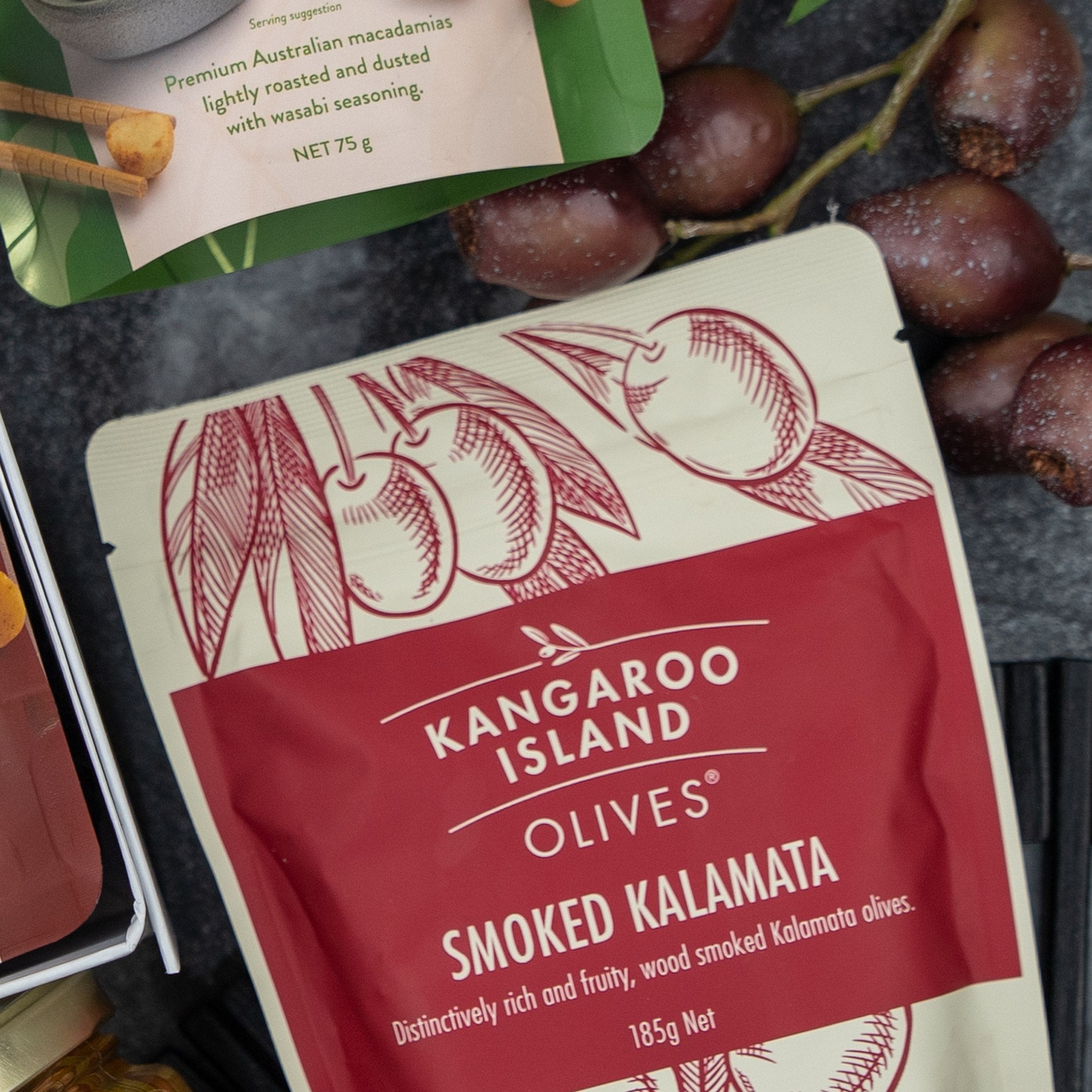 A close-up of The Hamper Boutique Co’s Flavour Fiesta Gift Hamper package next to fresh olives and other snacks on a dark surface.