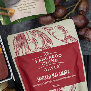 A close-up of The Hamper Boutique Co’s Flavour Fiesta Gift Hamper package next to fresh olives and other snacks on a dark surface.