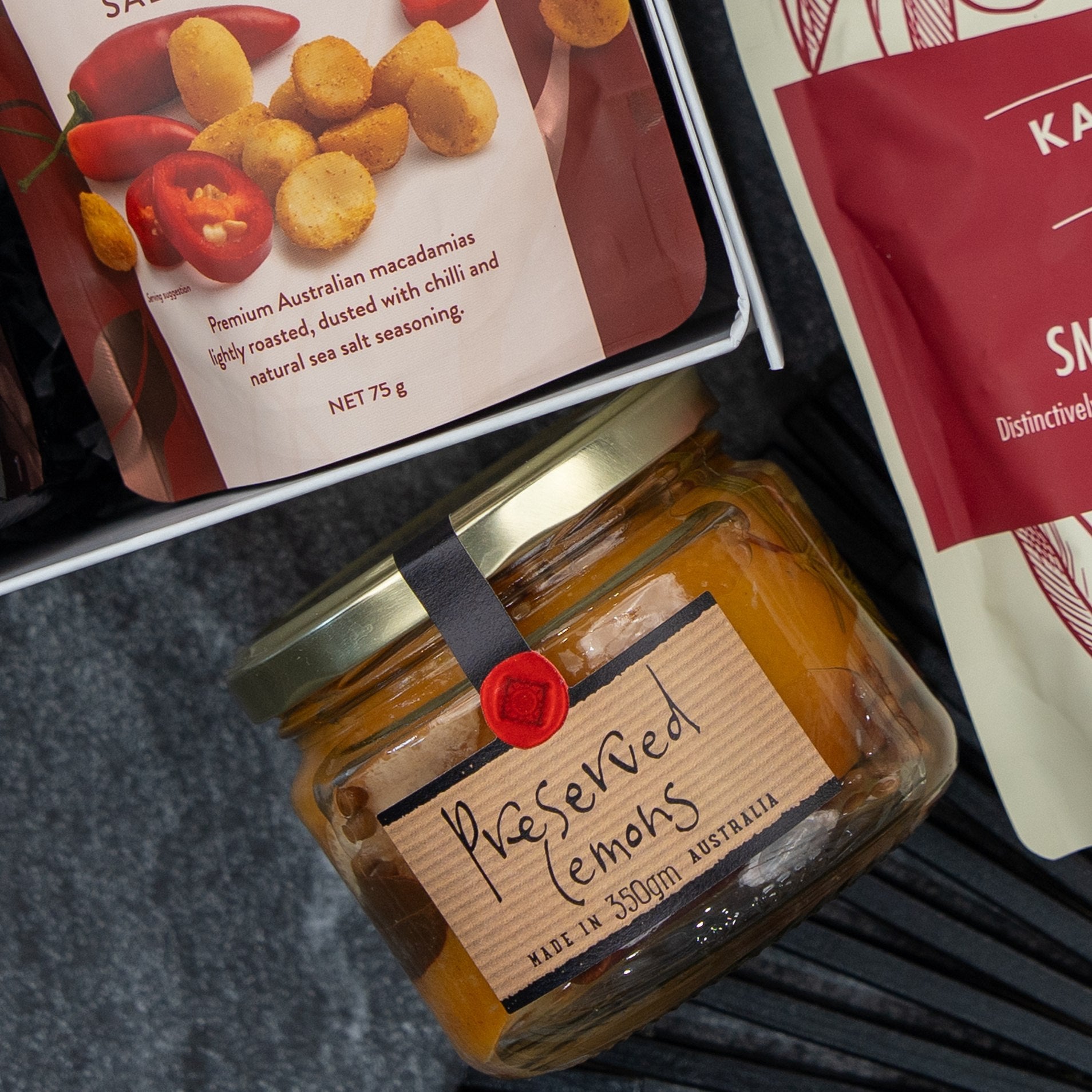 The Flavour Fiesta Gift Hamper by The Hamper Boutique Co features a jar of preserved lemons with a gold lid, alongside roasted macadamias and another snack, all neatly arranged together.