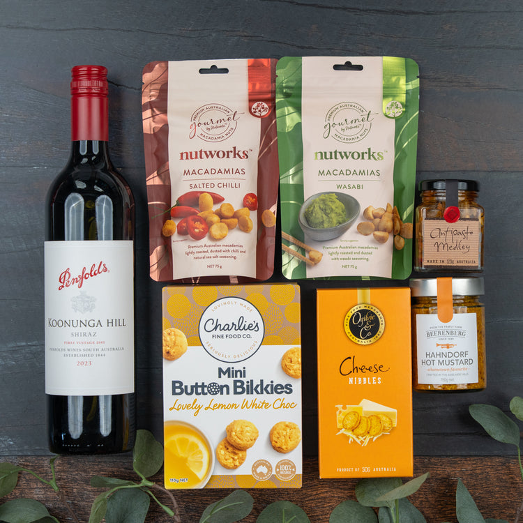 The Hamper Boutique Co Festive Treats Gift Hamper is displayed on dark wood and includes a bottle of Penfolds Shiraz, Nutworks macadamias, hot mustard, oat honey mini jar, Charlie’s lemon white choc bikkies & cheese nibbles.
