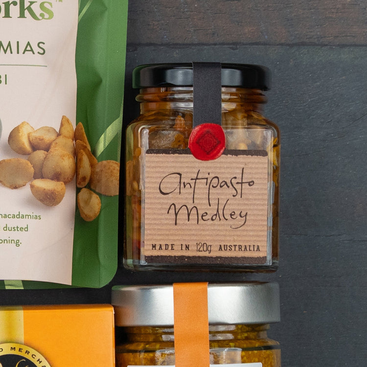 A Festive Treats Gift Hamper by The Hamper Boutique Co sits on a dark surface, featuring salted macadamias, an Antipasto Medley jar, and another jar with an orange ribbon partially visible.