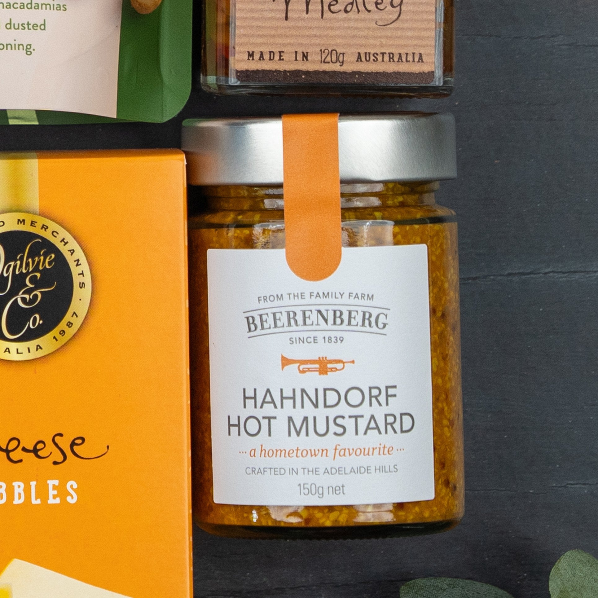 Close-up of The Hamper Boutique Co Festive Treats Gift Hamper, featuring gourmet items like Beerenberg Hahndorf Hot Mustard with orange label and ribbon, part of an Ogilvie & Co. cheese pack, plus a “Made in Australia” jar with a wooden lid.