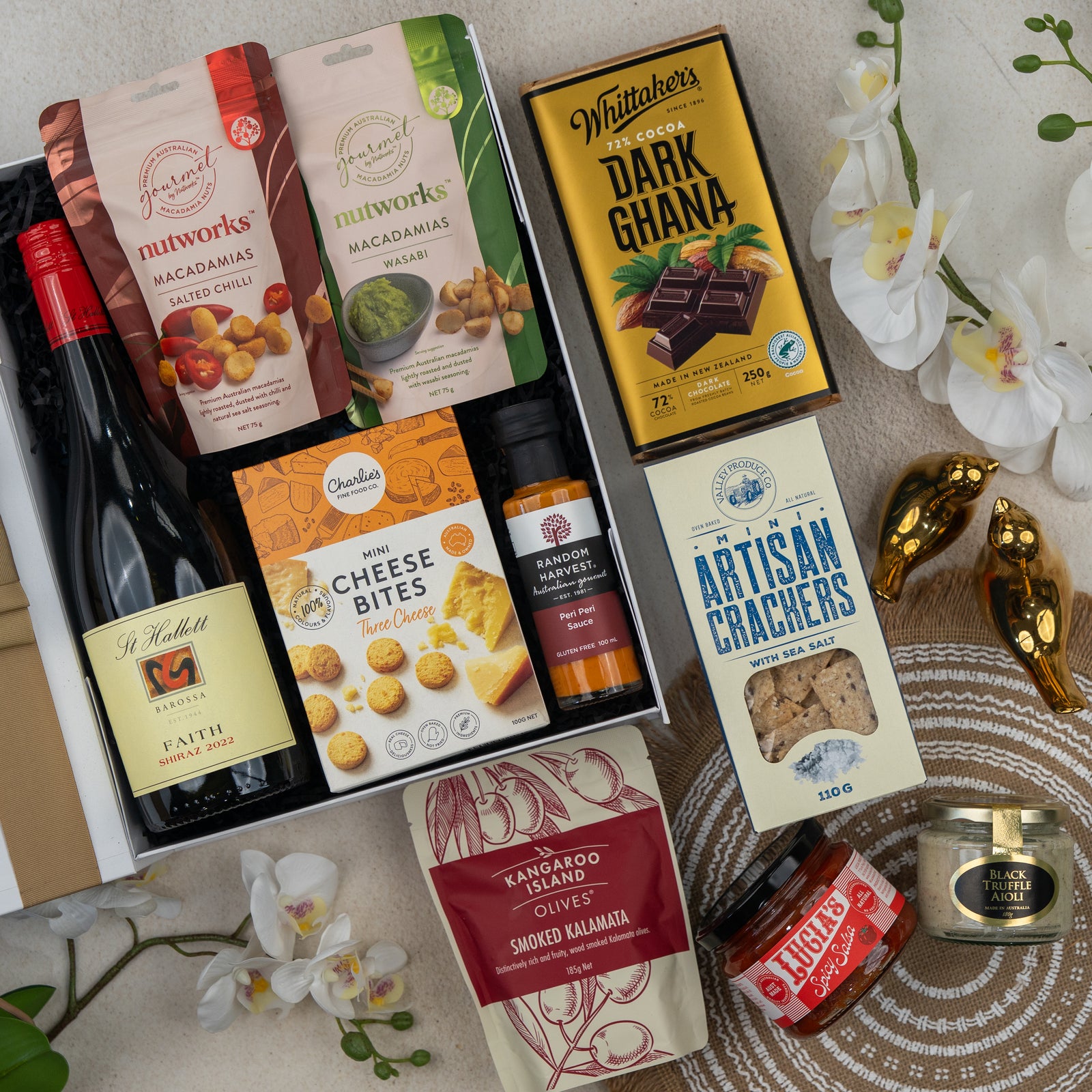 The Hamper Boutique Co’s Red Wine Gourmet Collection Hamper features wine, macadamias, dark chocolate, crackers, cheese bites, smoked kalamata olives, honey, relish, and a gold bird figurine on display with orchids.