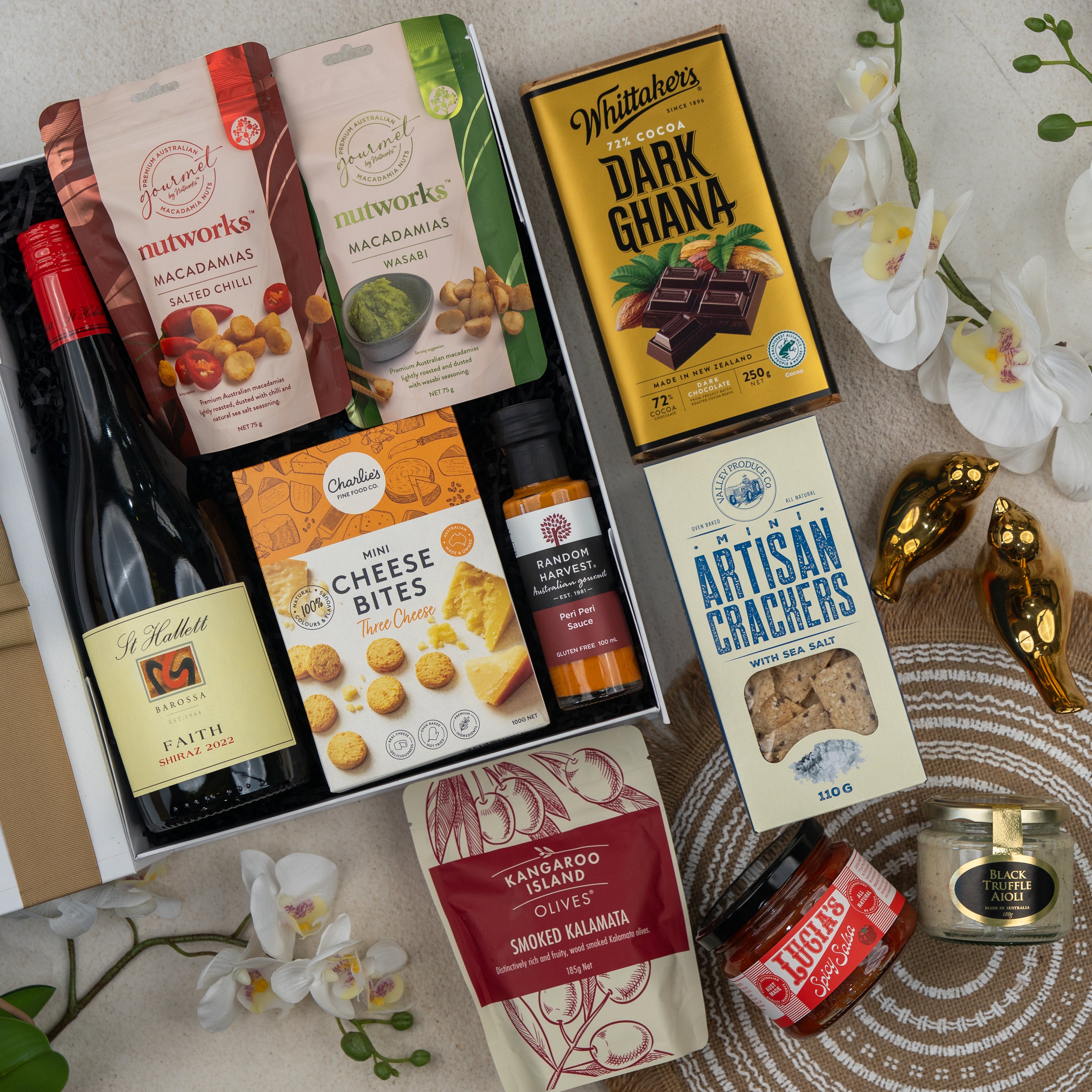 The Hamper Boutique Co’s Red Wine Gourmet Collection Hamper features wine, macadamias, dark chocolate, crackers, cheese bites, smoked kalamata olives, honey, relish, and a gold bird figurine on display with orchids.