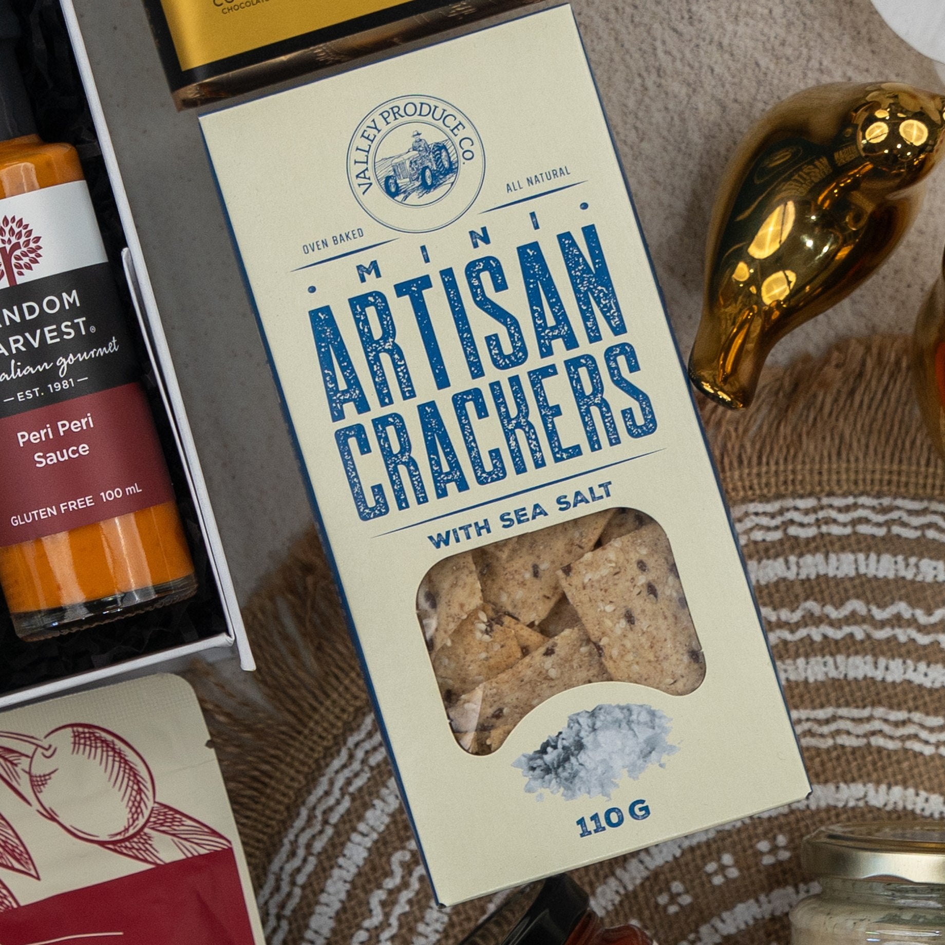 The Hamper Boutique Co Red Wine Gourmet Collection Hamper featuring Valley Produce Co. Mini Artisan Crackers with Sea Salt, Peri Peri sauce, a gold decorative piece, and part of a white basket.