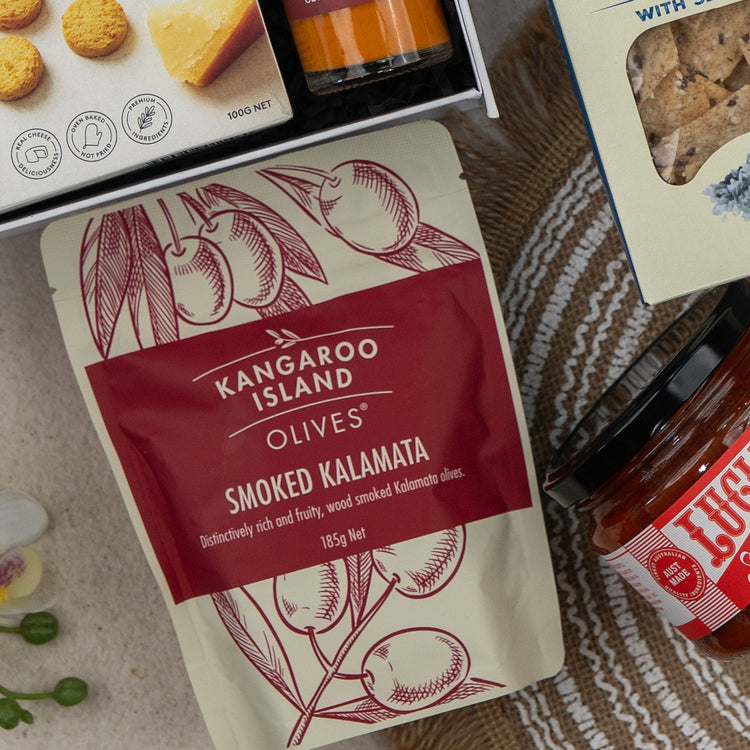 A top-down view of The Hamper Boutique Co Red Wine Gourmet Collection Hamper, featuring a red and beige pouch, crackers, cookies, and a jar of red sauce, all arranged on a woven mat.