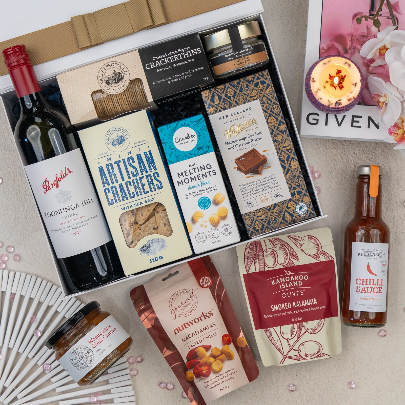 The Hamper Boutique Co’s Deluxe Grazing Gift Hamper features red wine, crackers, cookies, sauce, nuts, smoked olives, a candle, a fan, and a floral pink book arranged in and around a white gift box on a light surface.
