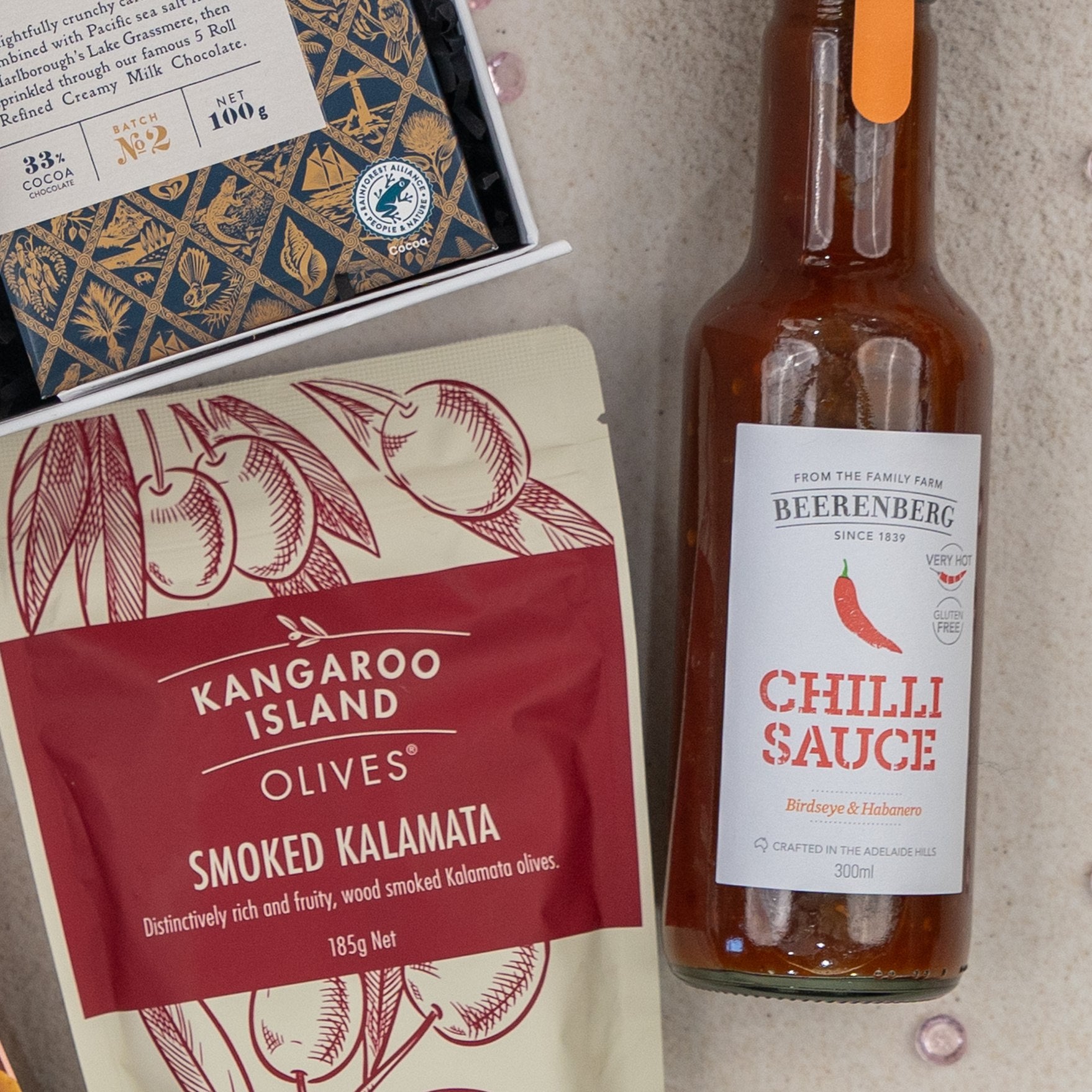 A flat lay of The Hamper Boutique Co Deluxe Grazing Gift Hamper, featuring gourmet items including chocolates, Kangaroo Island smoked Kalamata olives, and Beerenberg chilli sauce on a light surface.