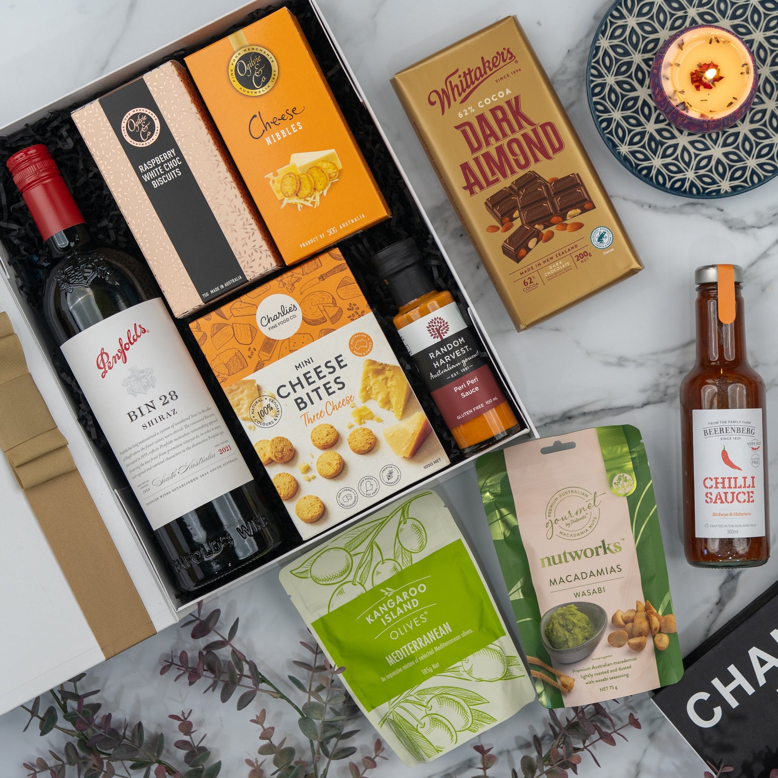 The Penfolds Celebration Hamper by The Hamper Boutique Co features red wine, cheese bites, crackers, nuts, dark almond chocolate, chili sauce, and a candle with greenery on a marble surface—perfect for gifting.