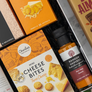A close-up of The Hamper Boutique Co's Penfolds Celebration Hamper, featuring orange-packaged cookies, mini cheese bites, a bottle of peri peri sauce, and part of a chocolate bar arranged on black shredded paper.