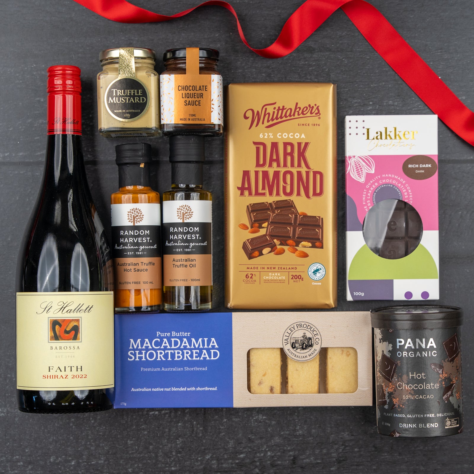 The Hamper Boutique Co's Luxury Indulgence Gift Hamper features wine, chocolates, shortbread, sauces, truffle mustard, dark almond chocolate, and hot chocolate, all beautifully arranged with a red ribbon on a dark surface.