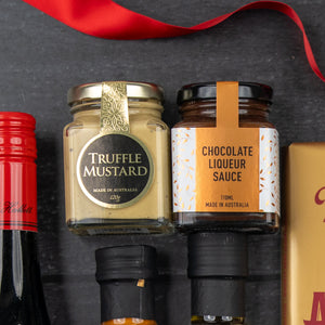 The Hamper Boutique Co's Luxury Indulgence Gift Hamper features truffle mustard and chocolate liqueur sauce jars arranged on a dark surface, with a red ribbon and a bottle adding to the elegant display.