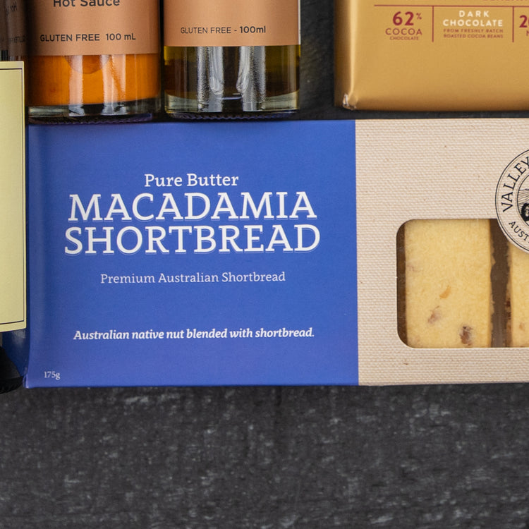 The Hamper Boutique Co’s Luxury Indulgence Gift Hamper features Pure Butter Macadamia Shortbread, dark chocolate, and hot sauce bottles, beautifully arranged on a dark surface.