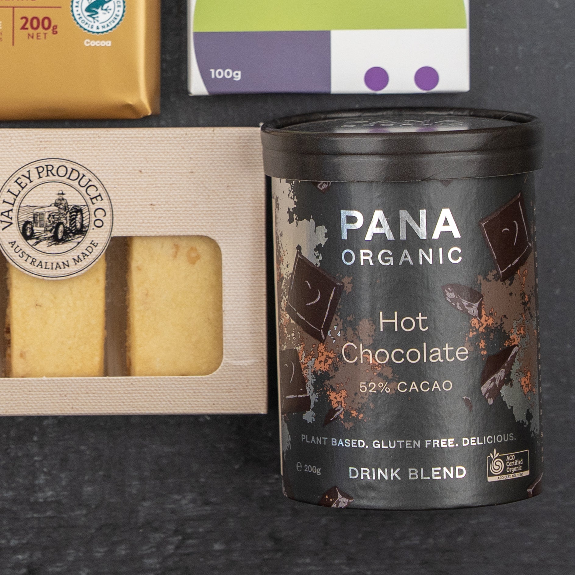 The Hamper Boutique Co Luxury Indulgence Gift Hamper features Pana Organic Hot Chocolate, shortbread cookies, a chocolate bar, and a package with purple and green accents, all elegantly arranged on a dark surface.