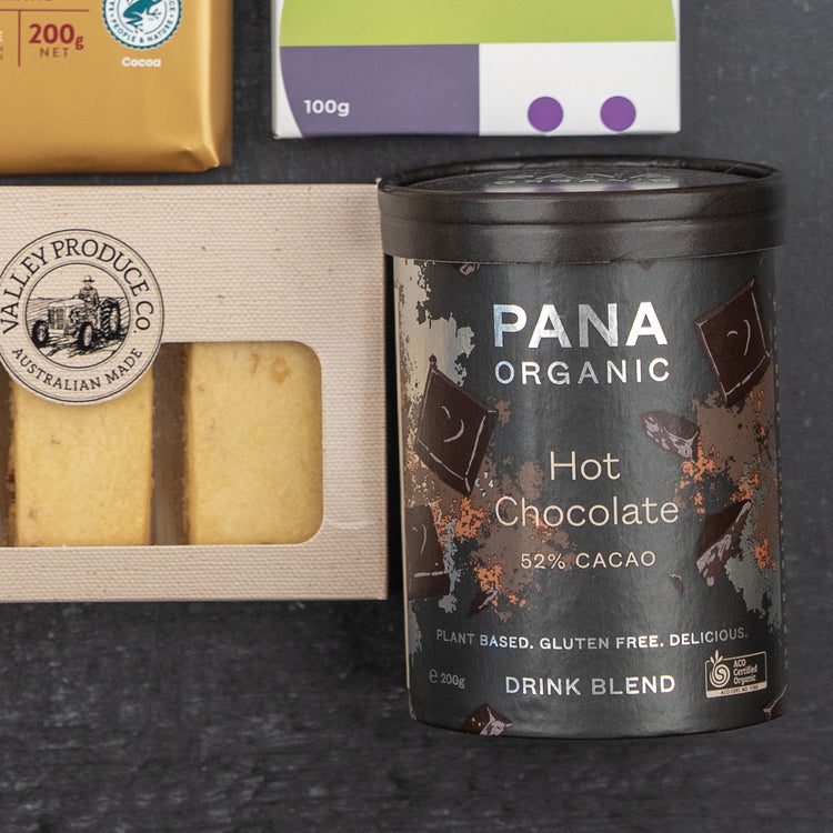 The Hamper Boutique Co Luxury Indulgence Gift Hamper features Pana Organic Hot Chocolate, shortbread cookies, a chocolate bar, and a package with purple and green accents, all elegantly arranged on a dark surface.