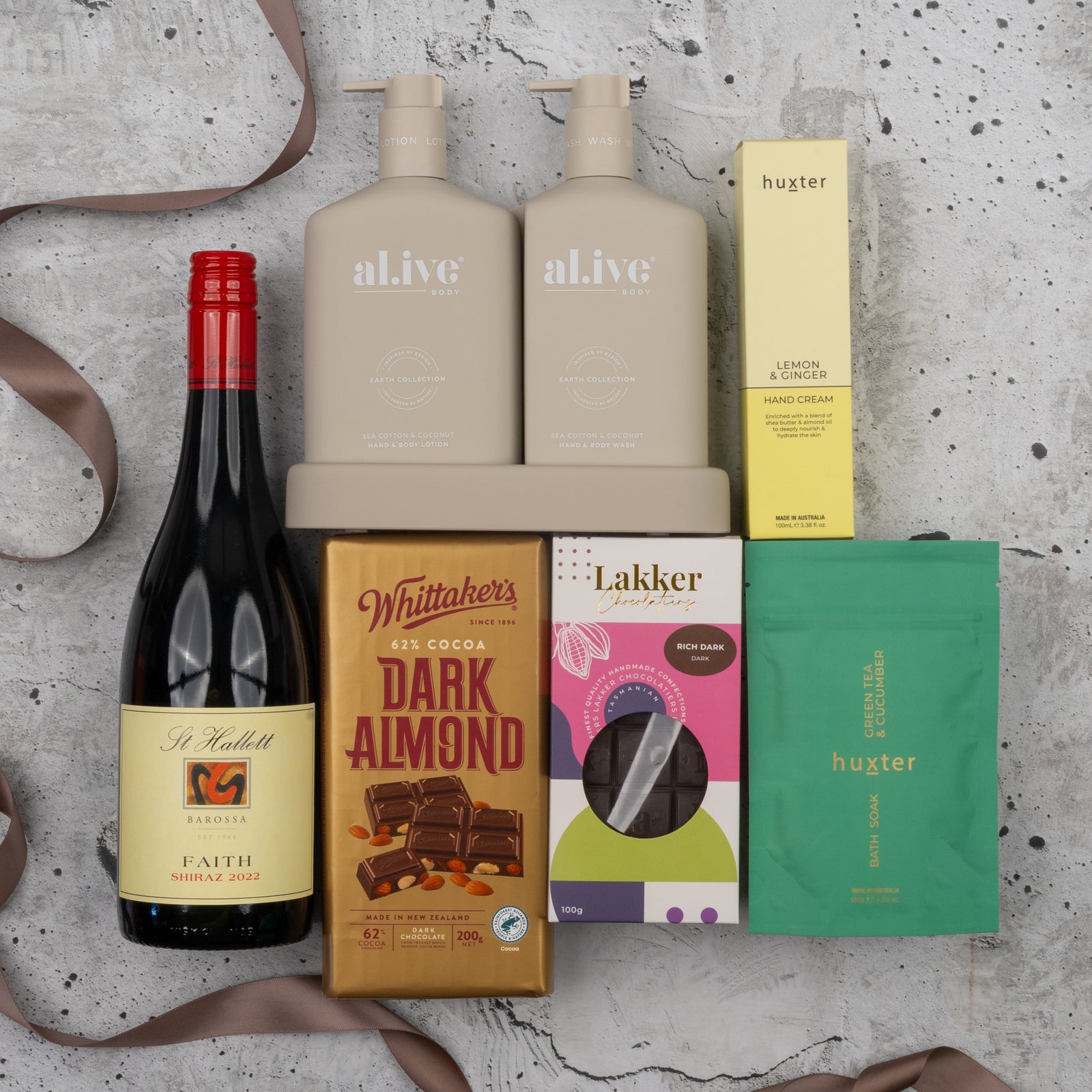 The Hamper Boutique Co's Indulgent Evening Escape includes St Hallett red wine, two beige al.ive body care bottles, Huxter hand cream, Whittakers dark almond chocolate, Lakker sweets, and Huxter green tea in a green pouch on a gray textured surface.