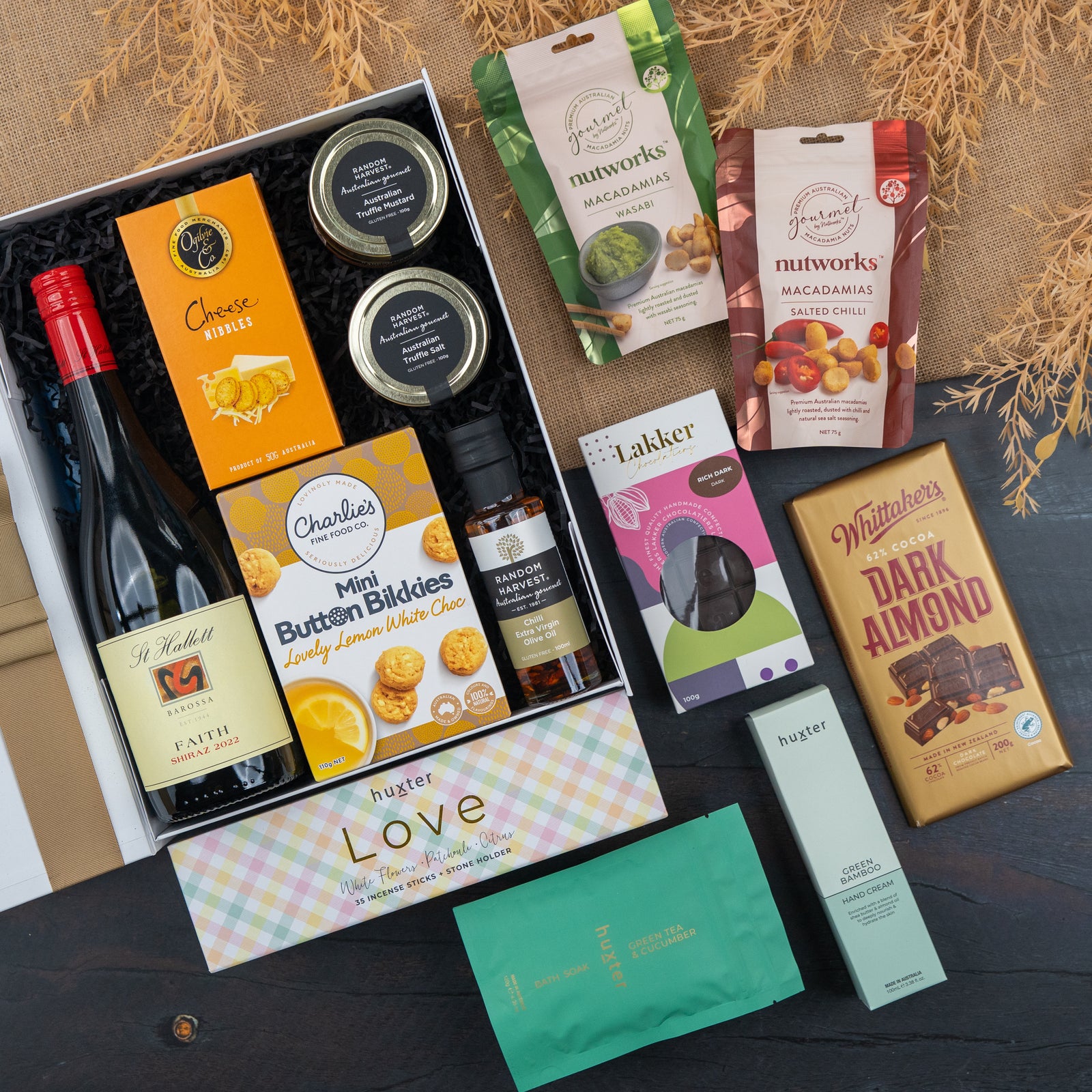 The Hamper Boutique Co's Red Wine Culinary Experience Hamper features red wine, cheese bites, crackers, spreads, chocolates, snacks, and tea—all beautifully arranged on a wooden surface with dried decorative plants in the background.