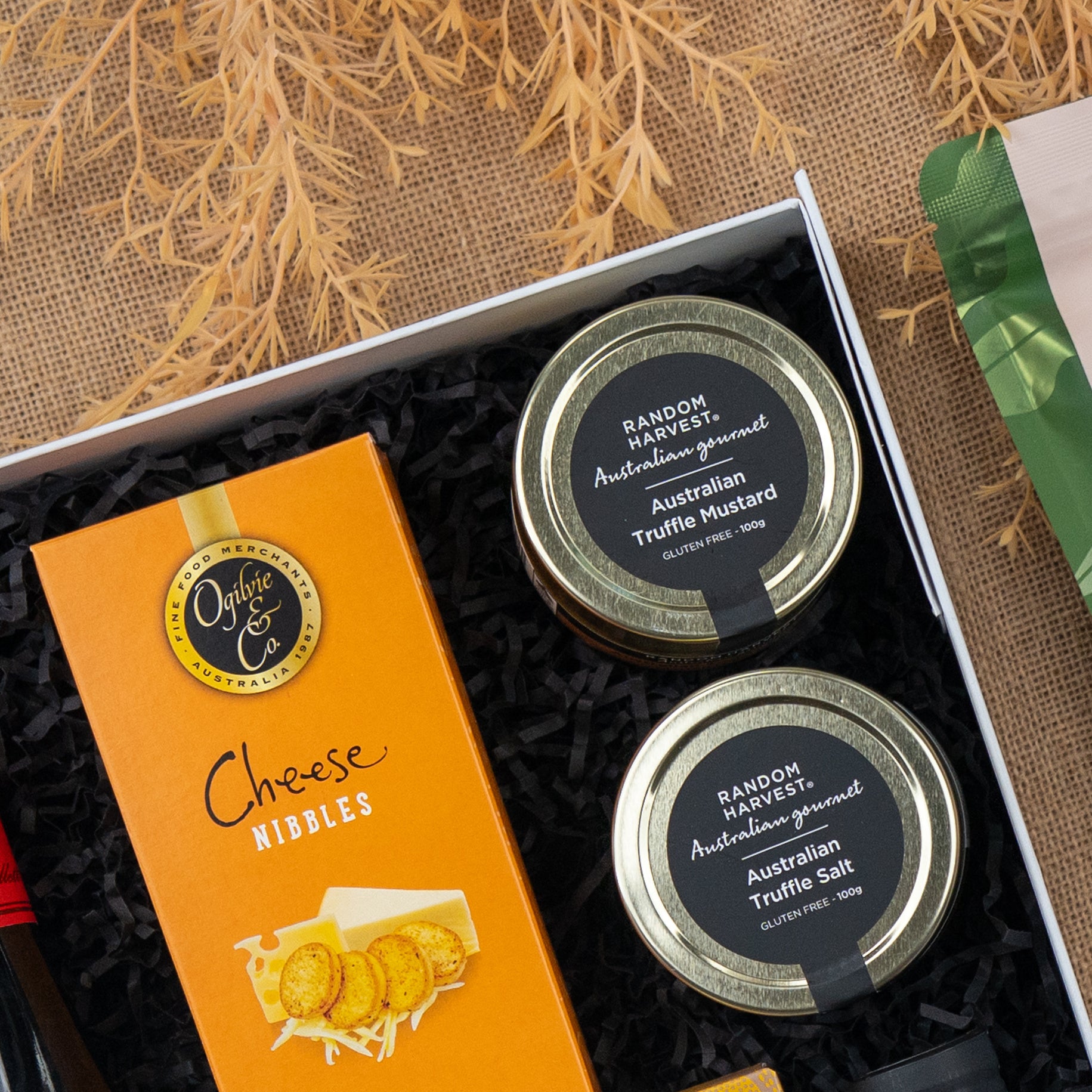 The Hamper Boutique Co's Red Wine Culinary Experience Hamper features cheese nibbles and jars of Australian Truffle Mustard and Truffle Salt, beautifully presented on black shredded paper with a rustic brown textured backdrop.