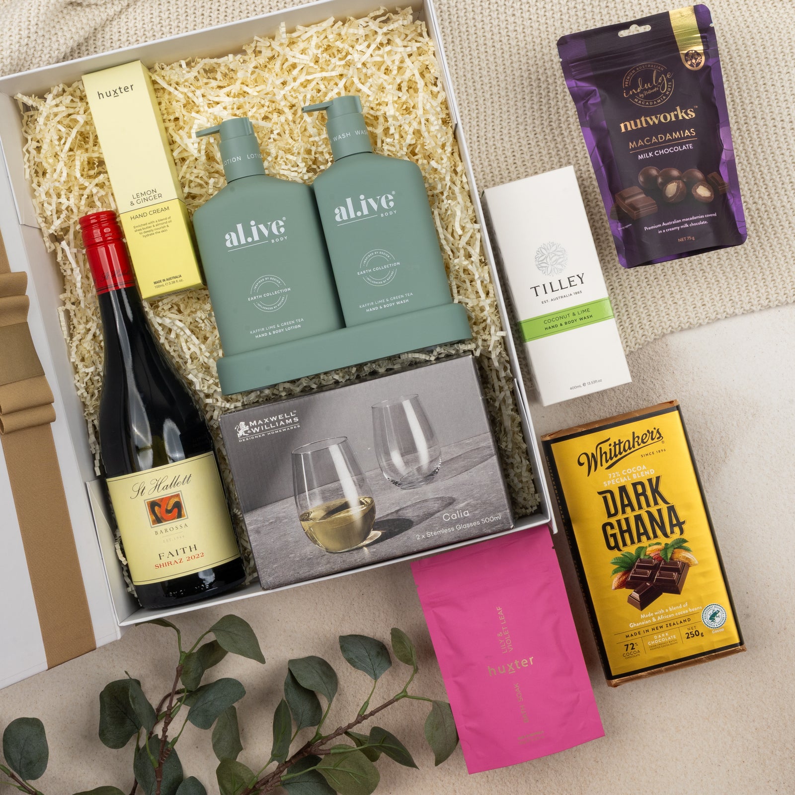 The Hamper Boutique Co's Relaxing Red Wine Hamper features wine, two green bottles, two wine glasses, snacks, chocolates including a gold-wrapped bar, hand cream, soap—all beautifully arranged with eucalyptus accents.