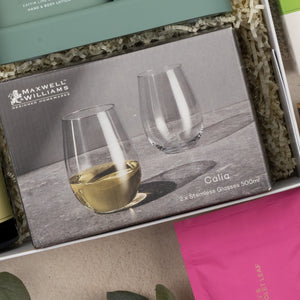 A close-up of The Hamper Boutique Co's Relaxing Red Wine Hamper shows two stemless glasses—one with white wine, one empty—in a gift box with shredded paper and other colorful packaged items.