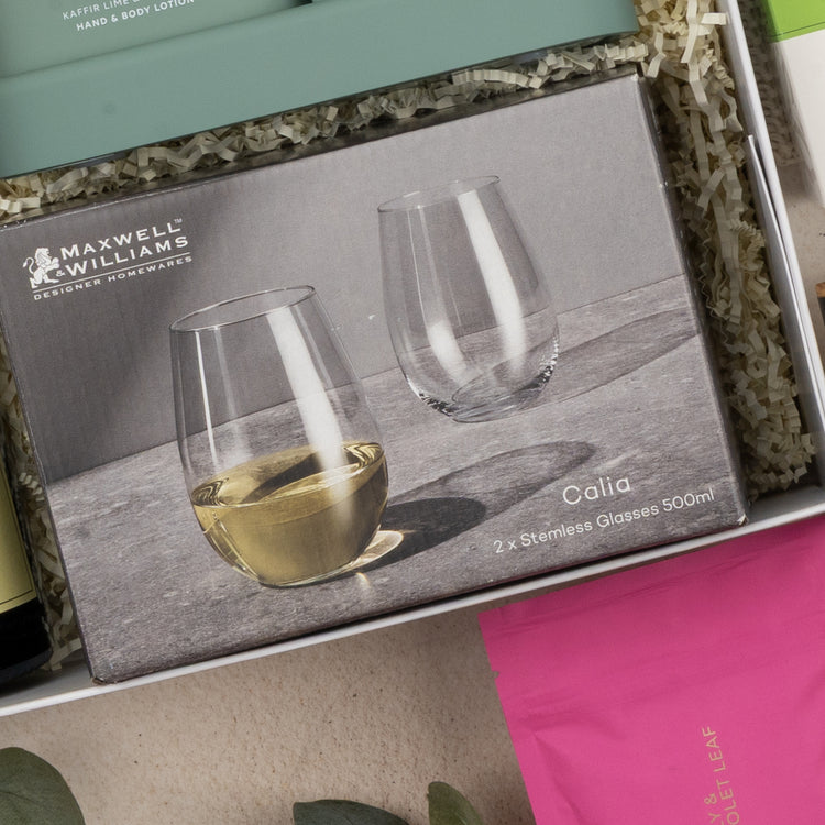 A close-up of The Hamper Boutique Co's Relaxing Red Wine Hamper shows two stemless glasses—one with white wine, one empty—in a gift box with shredded paper and other colorful packaged items.