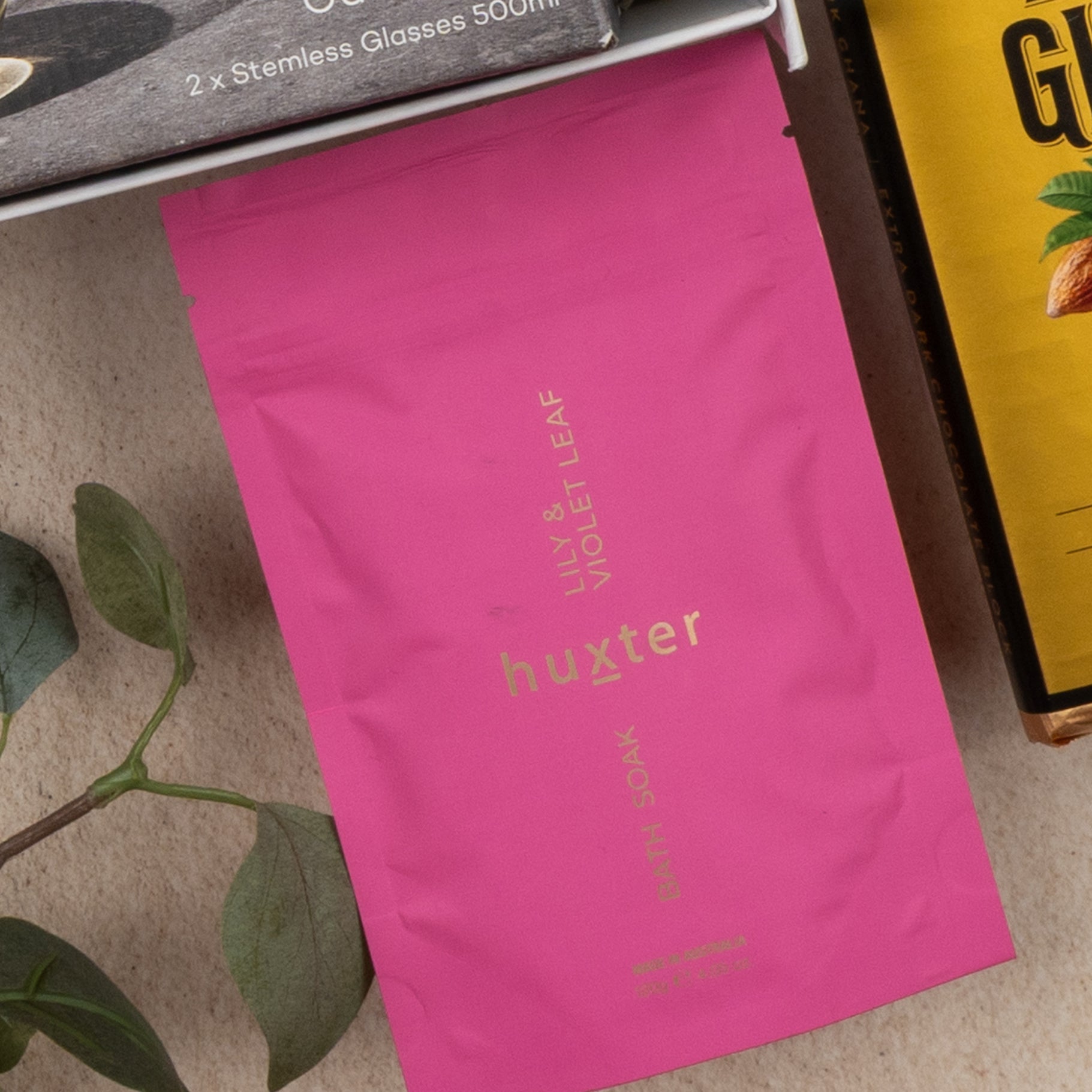 A Relaxing Red Wine Hamper from The Hamper Boutique Co sits on a surface with a pink huxter Lily & Violet Leaf Bath Soak, a plant branch, stemless glasses in a box, and a yellow package showing part of the word "GIFT.