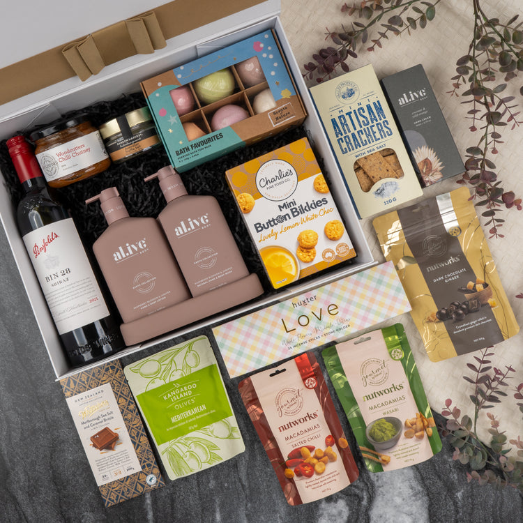 The Hamper Boutique Co's Deluxe Wine and Relaxation Hamper includes wine, biscuits, chocolates, nuts, tea, snacks, crackers, and bath products in a white ribboned box with decorative leaves on a marble surface.