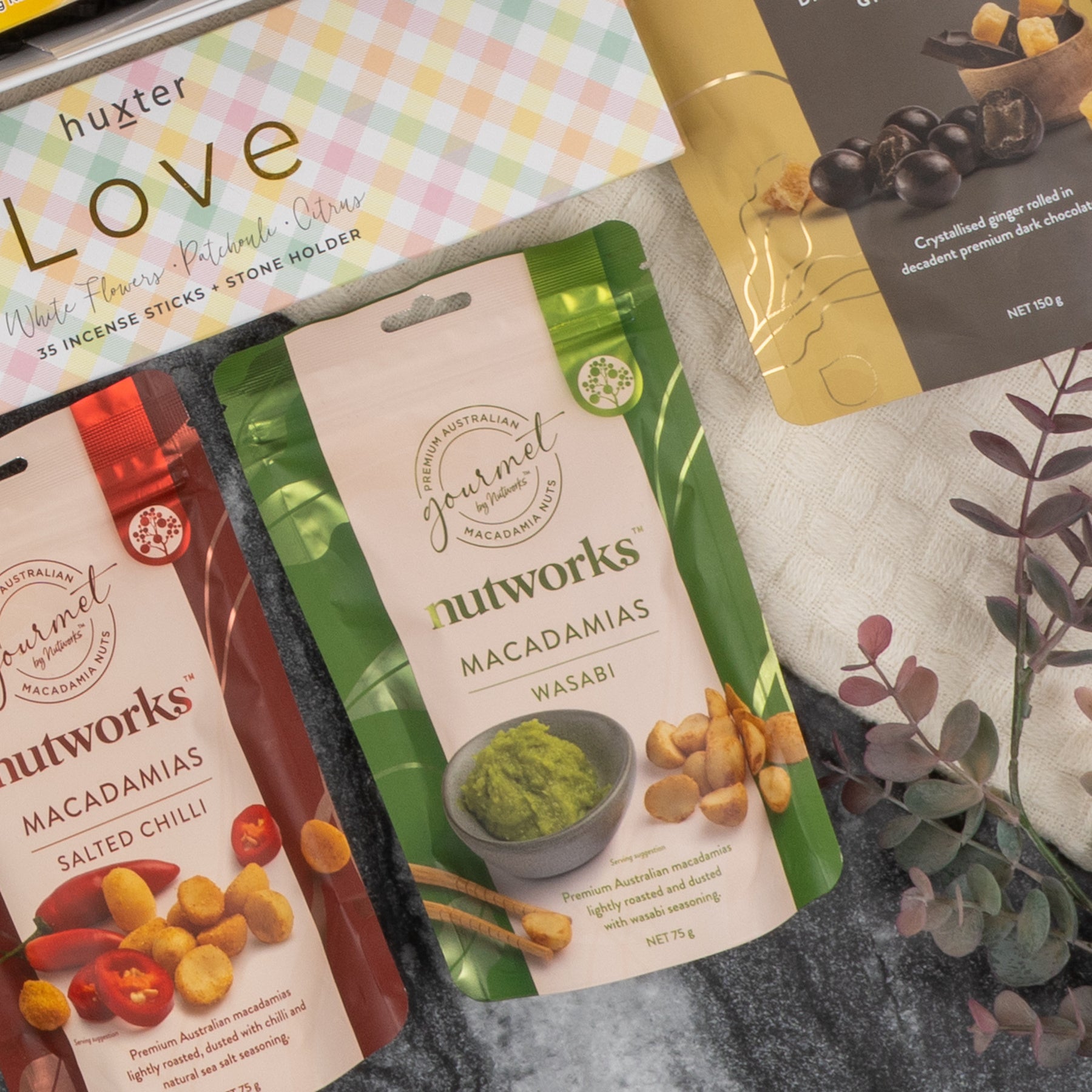 The Deluxe Wine and Relaxation Hamper by The Hamper Boutique Co features assorted snack packs, including Nutworks macadamias in salted chili and wasabi flavors, arranged on a gray surface with a leafy branch and a pastel huxter Love box.