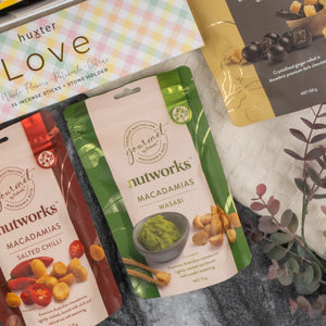 The Deluxe Wine and Relaxation Hamper by The Hamper Boutique Co features assorted snack packs, including Nutworks macadamias in salted chili and wasabi flavors, arranged on a gray surface with a leafy branch and a pastel huxter Love box.