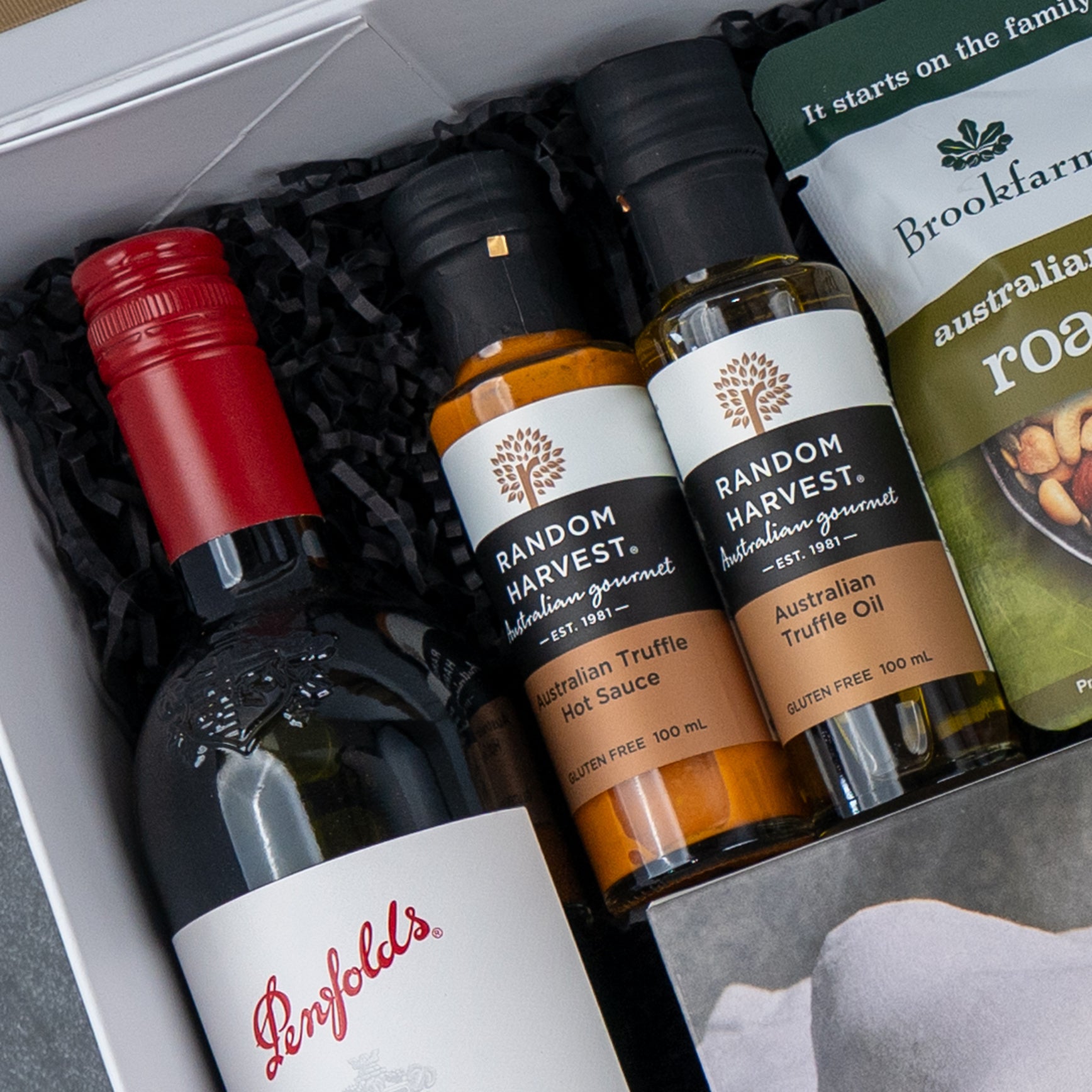 A close-up of The Hamper Boutique Co’s Luxe Dining Red Wine Hamper with Penfolds wine, two Random Harvest Truffle condiments, and Brookfarm Australian roasted nuts on black shredded paper.