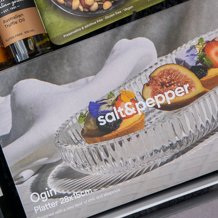 A close-up of a Luxe Dining Red Wine Hamper box by The Hamper Boutique Co, showing the hamper with gourmet appetizers like figs and edible flowers. Part of a truffle oil bottle appears above the box.
