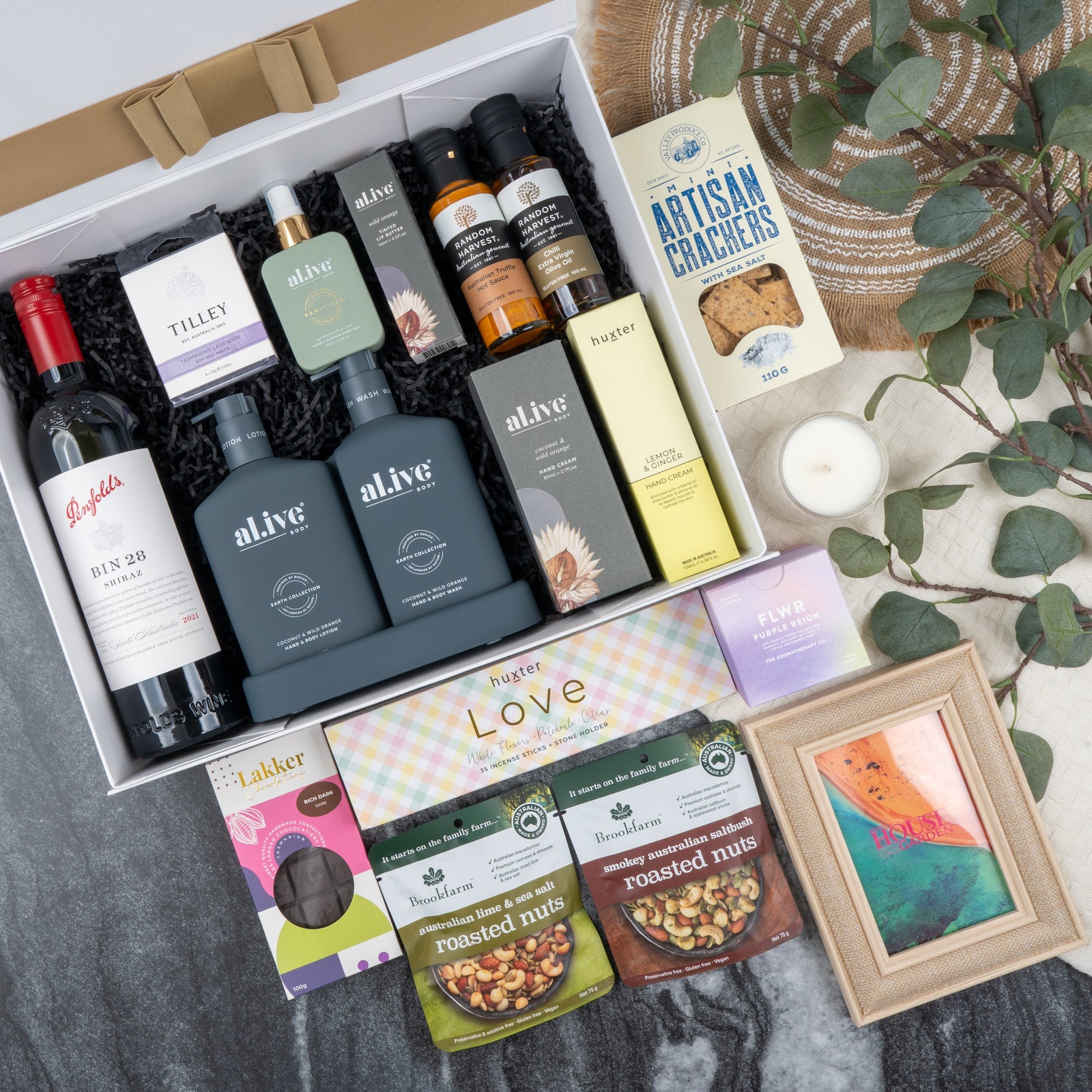 The Hamper Boutique Co’s Ultimate Red Wine Celebration Hamper features wine, skincare, crackers, chocolates, roasted nuts, oils, a candle, soap, and a framed picture—beautifully presented with leafy accents on marble and woven textures.