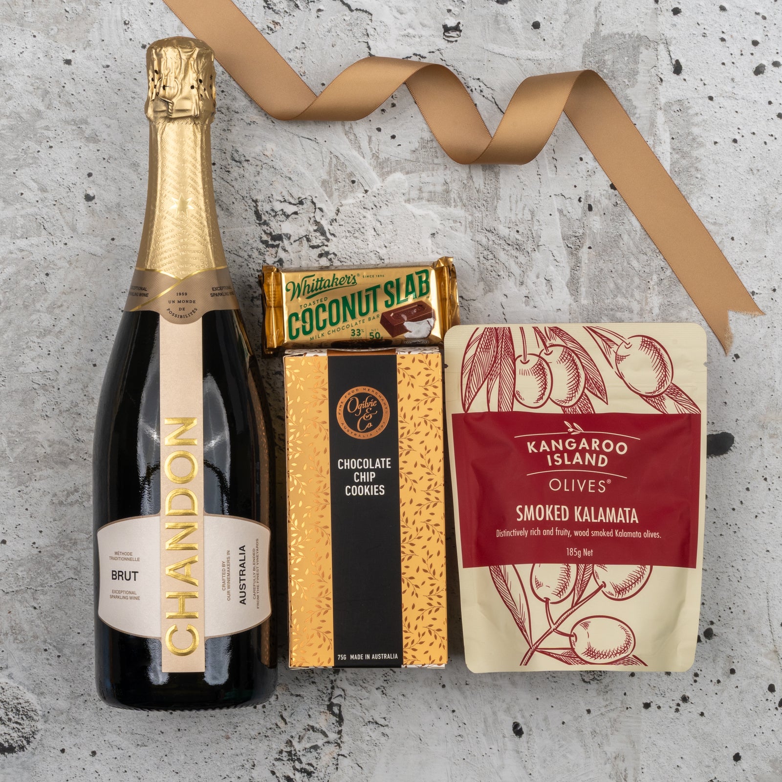 The Hamper Boutique Co's Chandon Sparkling Delights Hamper, featuring Chandon Brut, Whittaker’s coconut slab, choc chip cookies, and Kangaroo Island smoked Kalamata olives, is arranged on concrete with a gold ribbon.