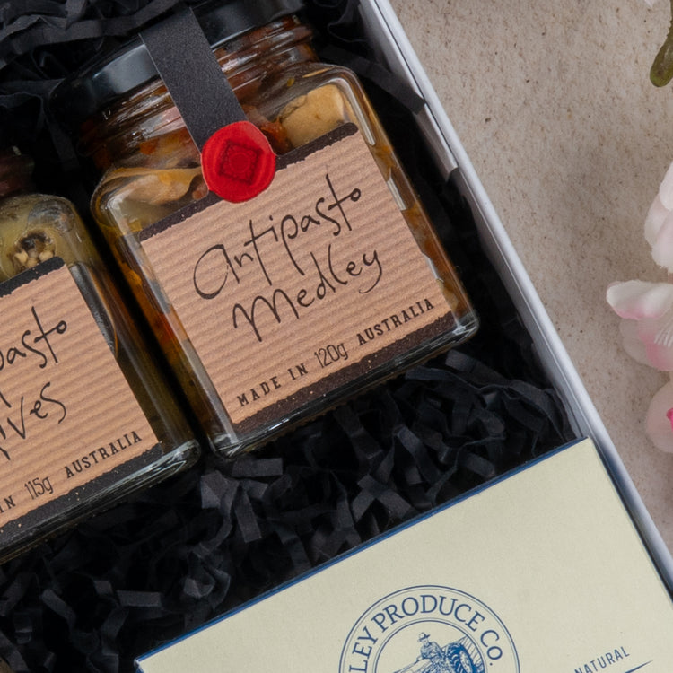 Two glass jars from The Hamper Boutique Co’s Chandon Celebration Hamper rest in black shredded paper. A cream envelope with a blue seal is at the bottom, while pink flowers flourish on the right.