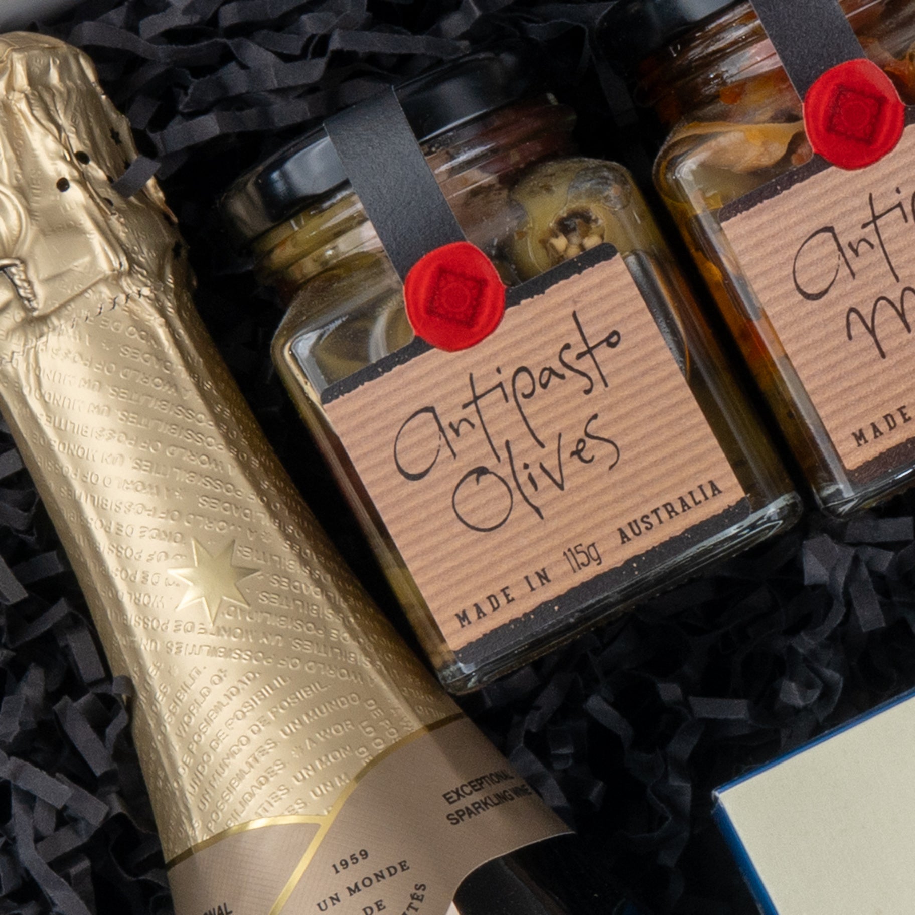 Close-up of The Hamper Boutique Co’s Chandon Celebration Hamper featuring a bottle of sparkling wine and jars of antipasto olives, all nestled in black shredded paper.