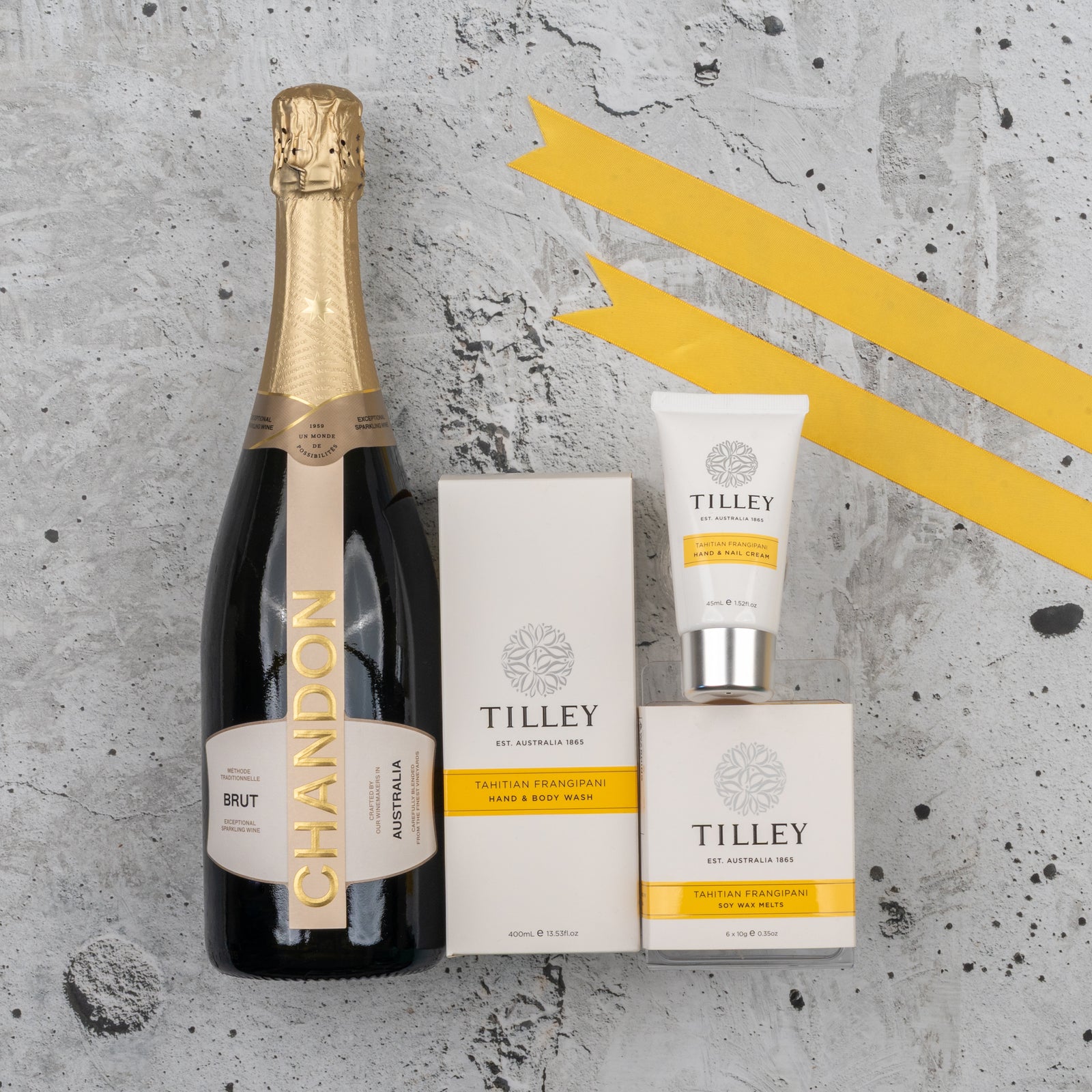 The Chandon Bubbles and Treats Hamper by The Hamper Boutique Co, featuring a bottle of Chandon Brut alongside three Tilley Tahitian Frangipani products, is elegantly arranged on a textured gray surface, with two yellow diagonal stripes highlighting the corner.