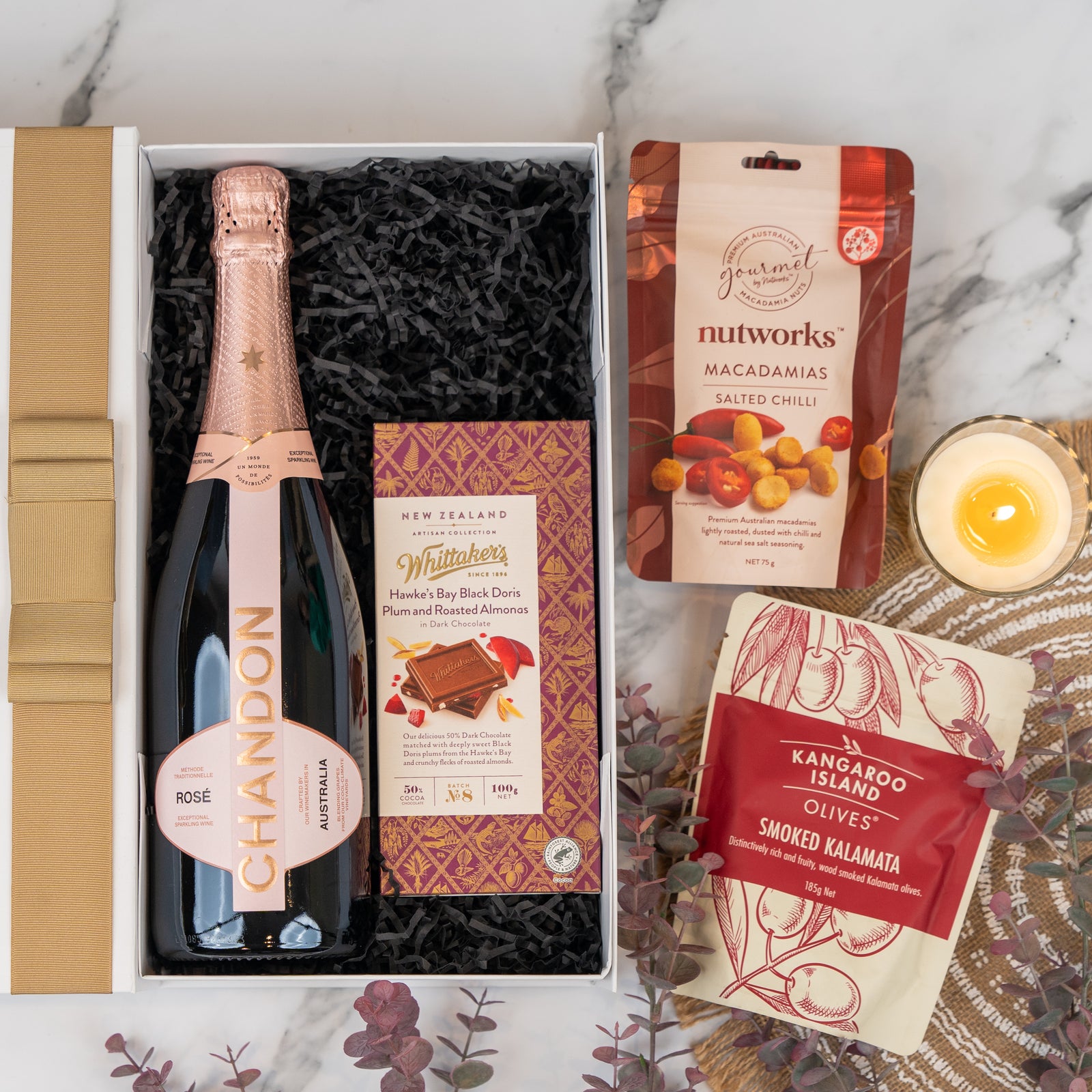 The Hamper Boutique Co’s Chandon Rosé Celebration Hamper features Chandon rosé, Whittaker’s chocolate, Nutworks salted chili macadamias, smoked Kalamata olives, a candle, and eucalyptus sprigs on marble.