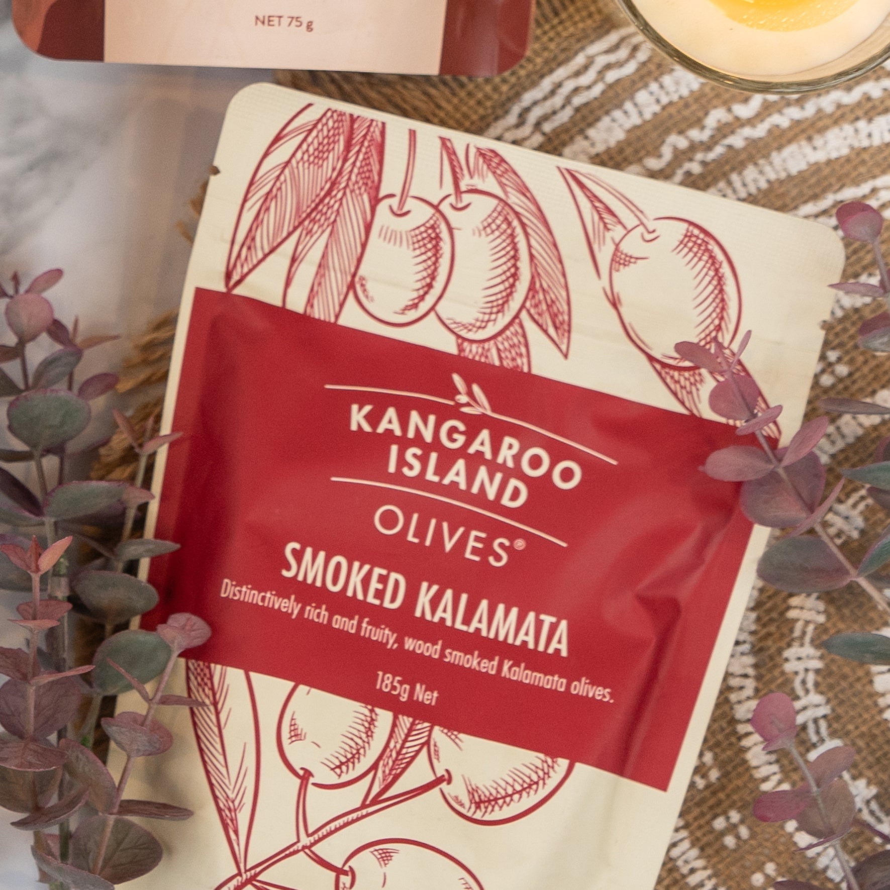 A box of The Hamper Boutique Co Chandon Rosé Celebration Hamper rests on a woven mat beside some eucalyptus leaves. The packaging is elegant, featuring cream and red tones with festive illustrations.